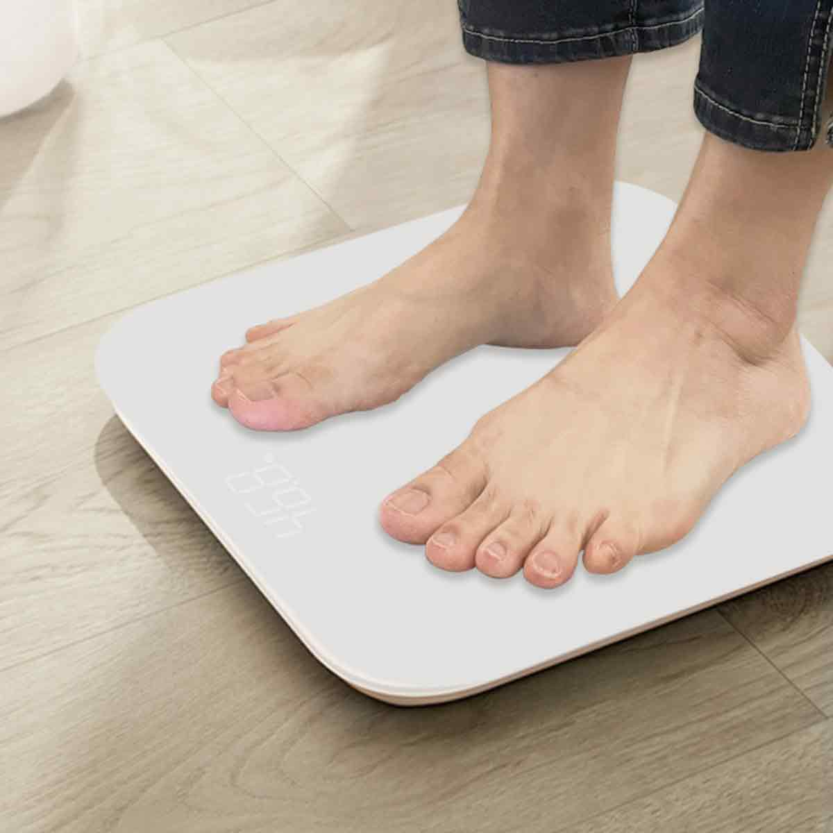 Barfuß auf einer weißen Waage, die 46,8 anzeigt. Jeans sind oben sichtbar. Der Boden ist aus Holz.