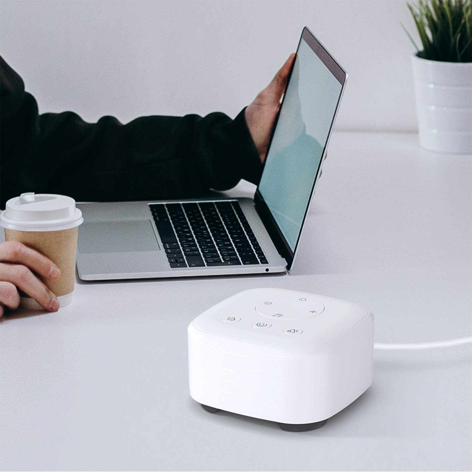 Eine weiße Klangmaschine, ein Laptop und eine Kaffeetasse auf einem Schreibtisch. Eine Person hält die Tasse und benutzt den Laptop.
