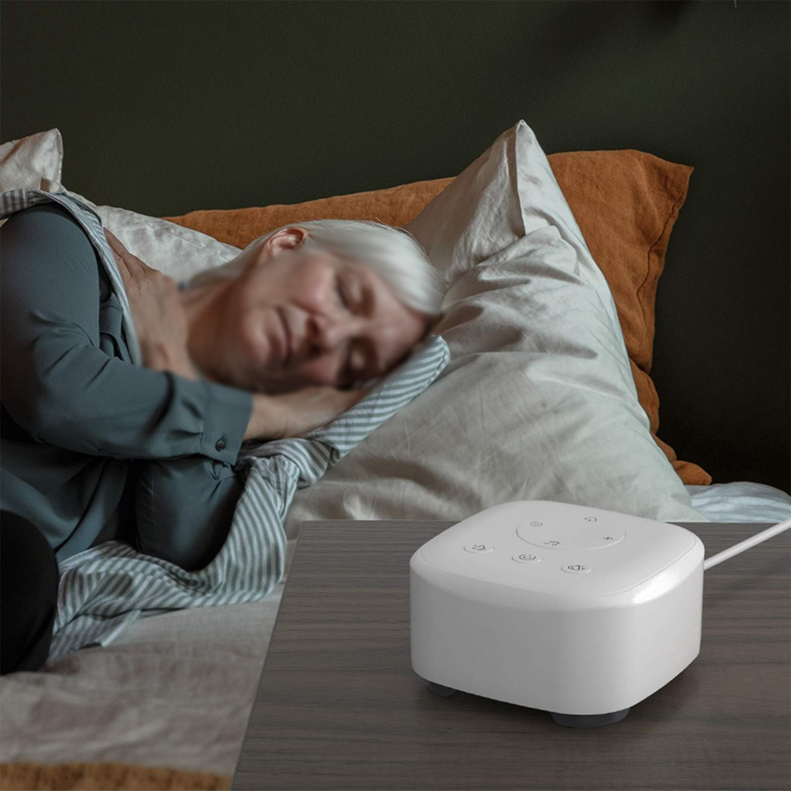 Eine Person schläft im Bett mit einer weißen Geräuschmaschine auf einem Nachttisch. Hinter dem Kopf der Person befinden sich Kissen.