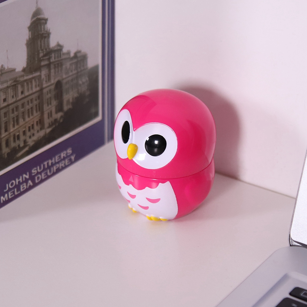 Ein rosa eulenformiger Behälter steht auf einer weißen Oberfläche. Links ist ein Buch mit einem Gebäude. Im Hintergrund ein Laptop.