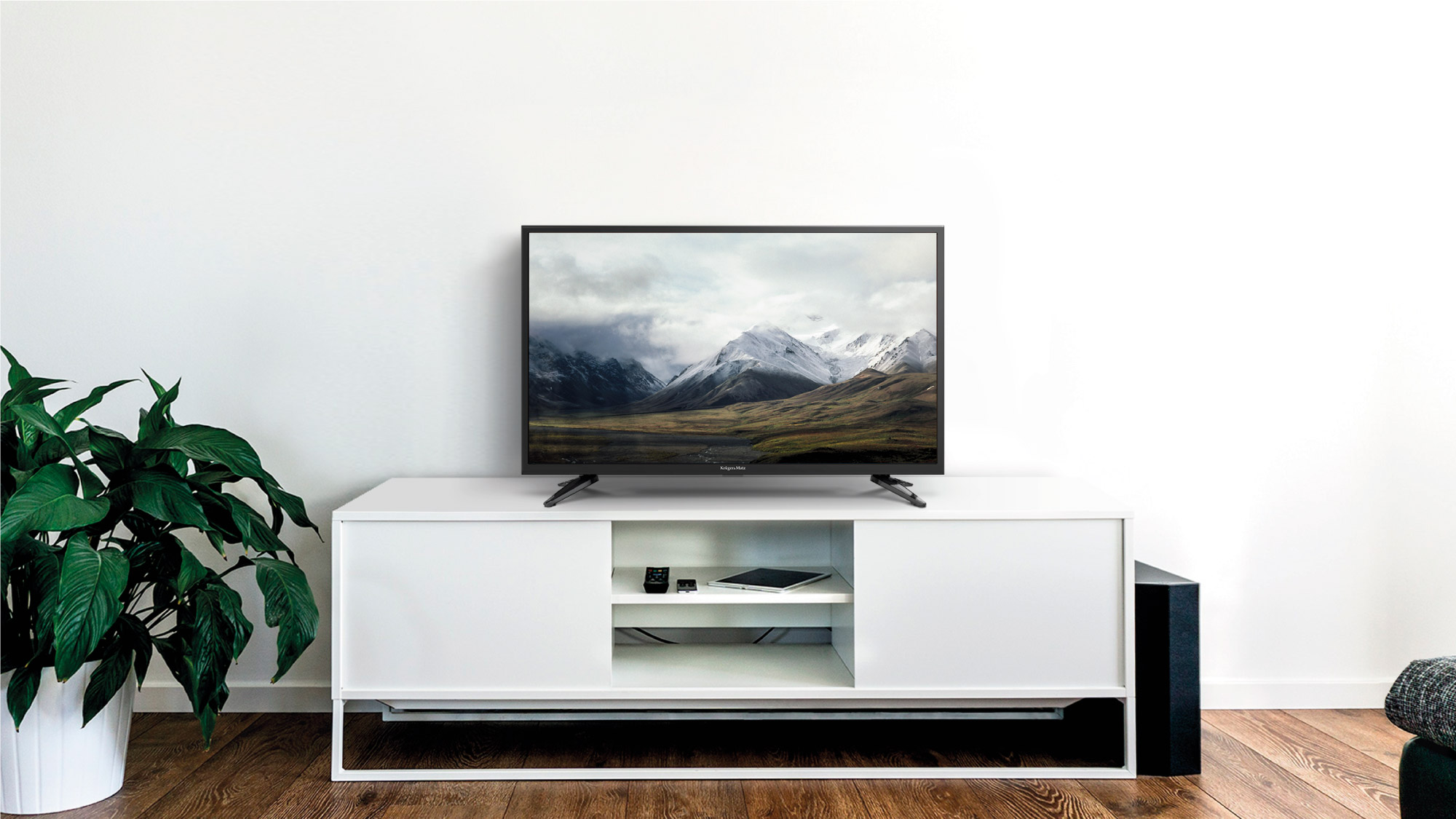 Telewizor wyświetla krajobraz górski. Stoi na białej szafce z rośliną i głośnikiem. Tłem jest biała ściana.