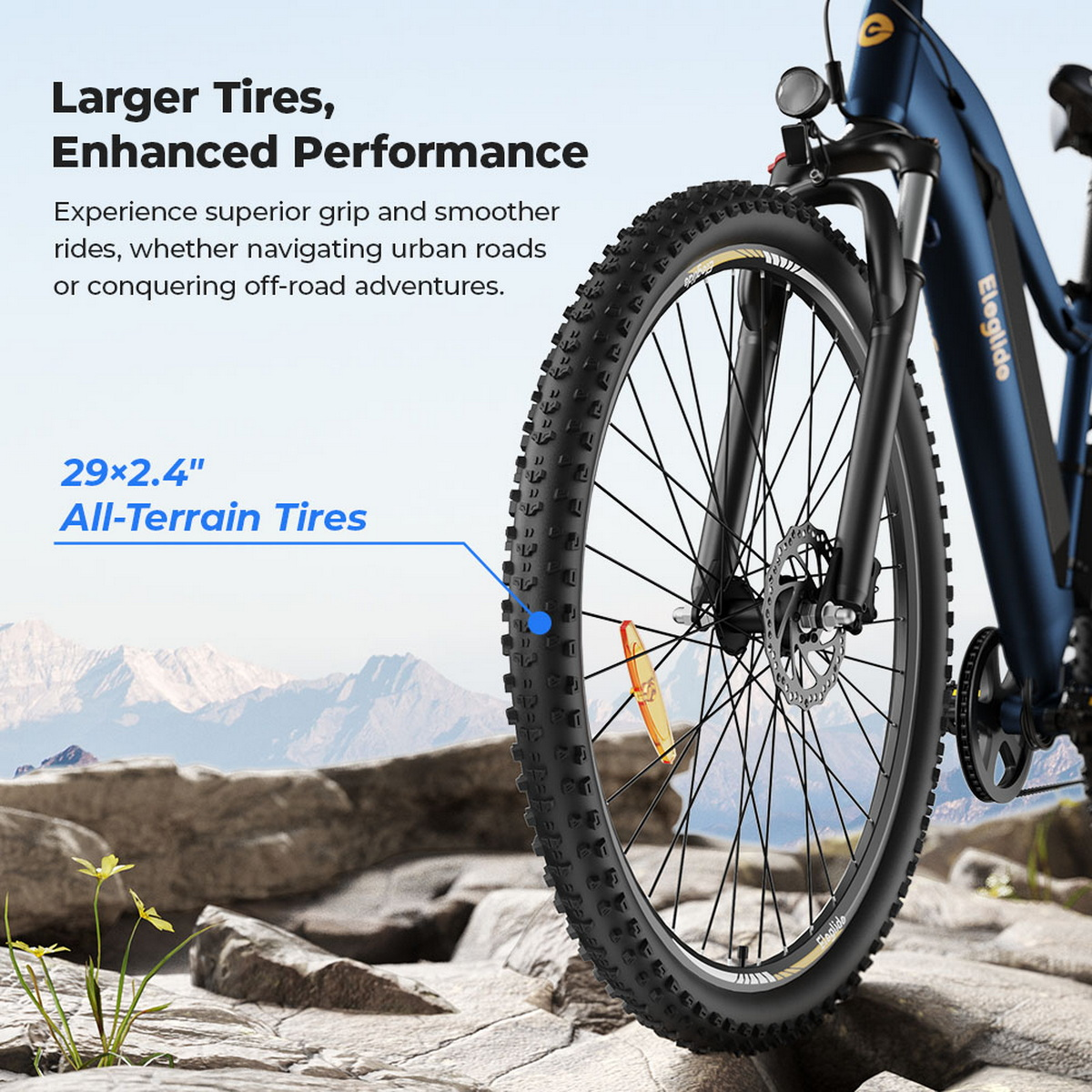 Nahaufnahme eines E-Mountainbike-Reifens. Der Reifen ist schwarz mit einem robusten Profil. Blauer Fahrradrahmen, felsiger Boden und Berge im Hintergrund.