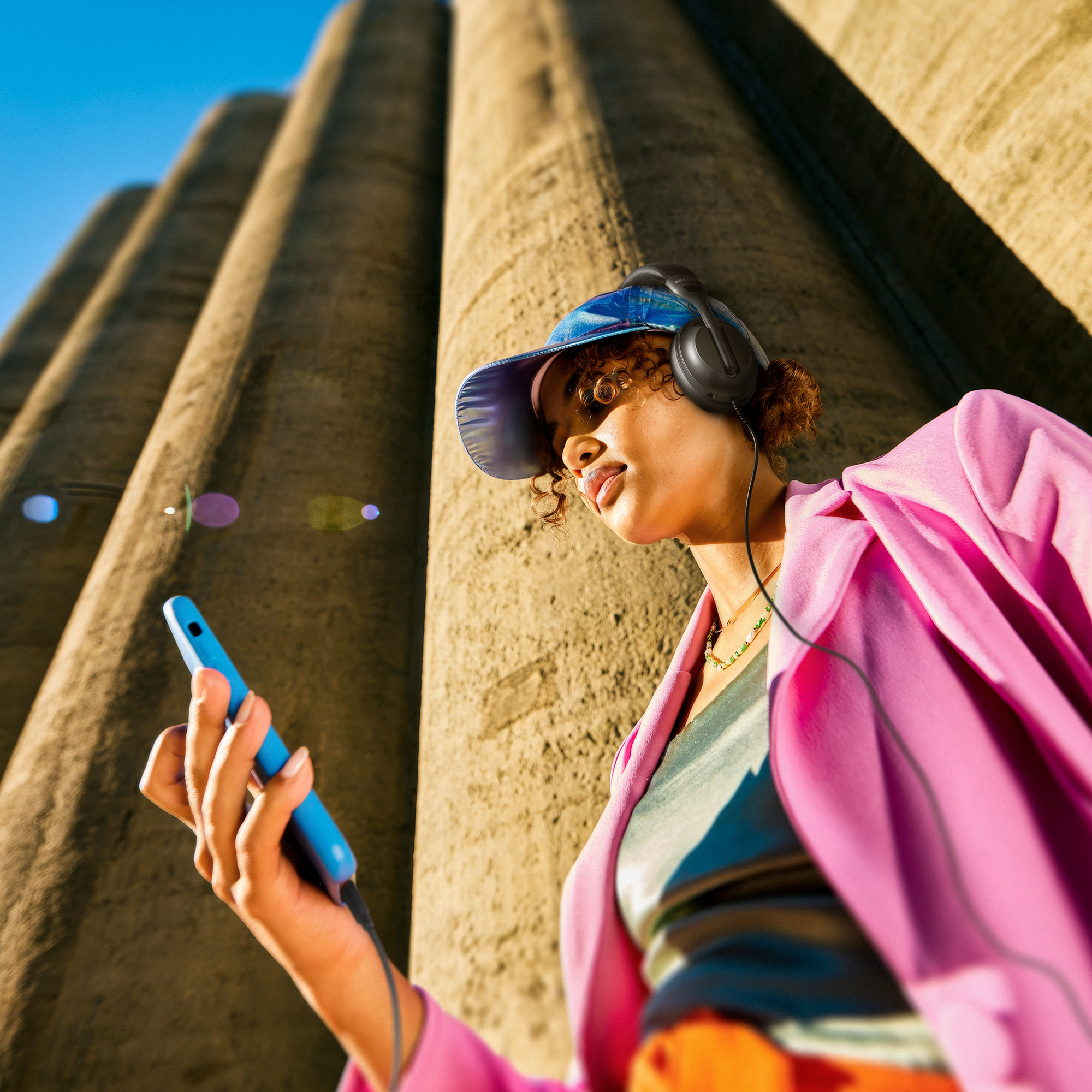 Kobieta ze słuchawkami i telefonem. W różowej kurtce i niebieskiej czapce, przed cylindrycznymi strukturami.