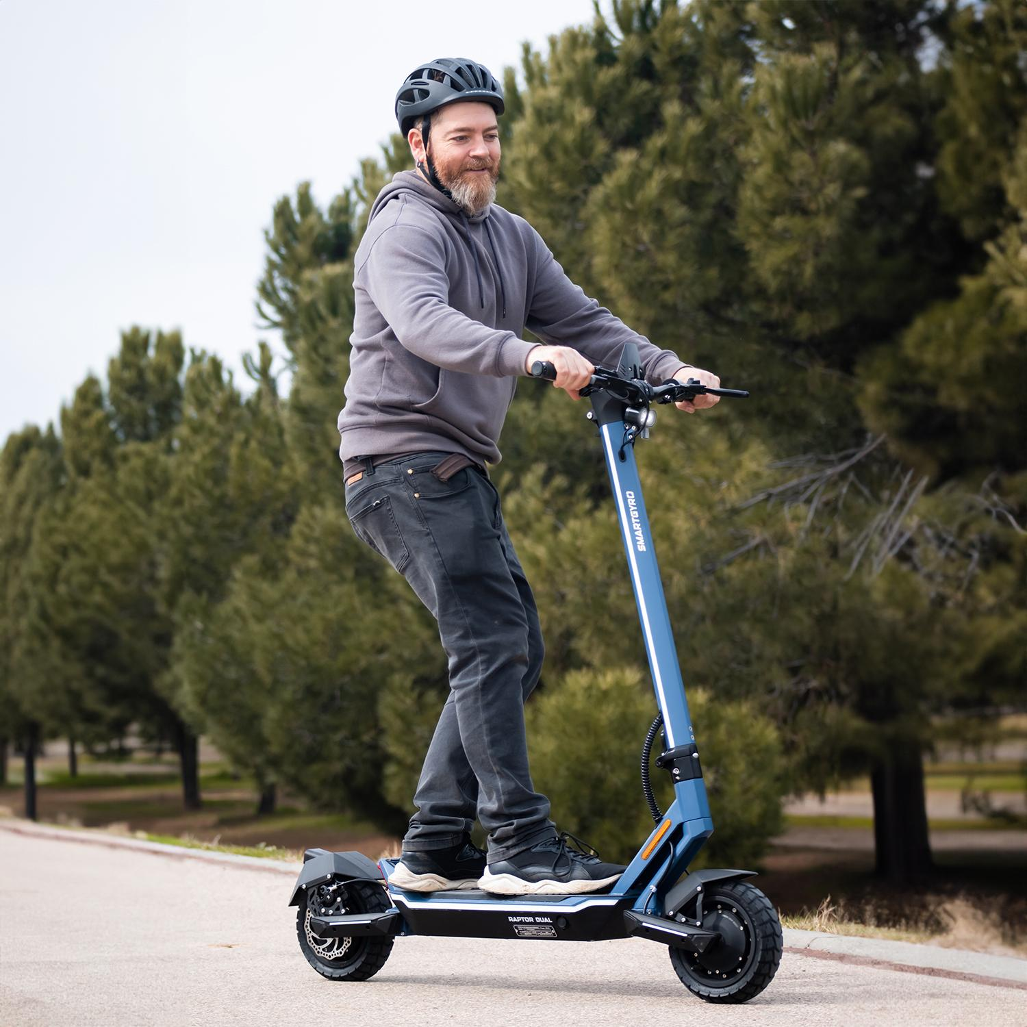 Un hombre monta un scooter eléctrico azul en una carretera pavimentada. Lleva casco y sudadera gris. Hay árboles al fondo.