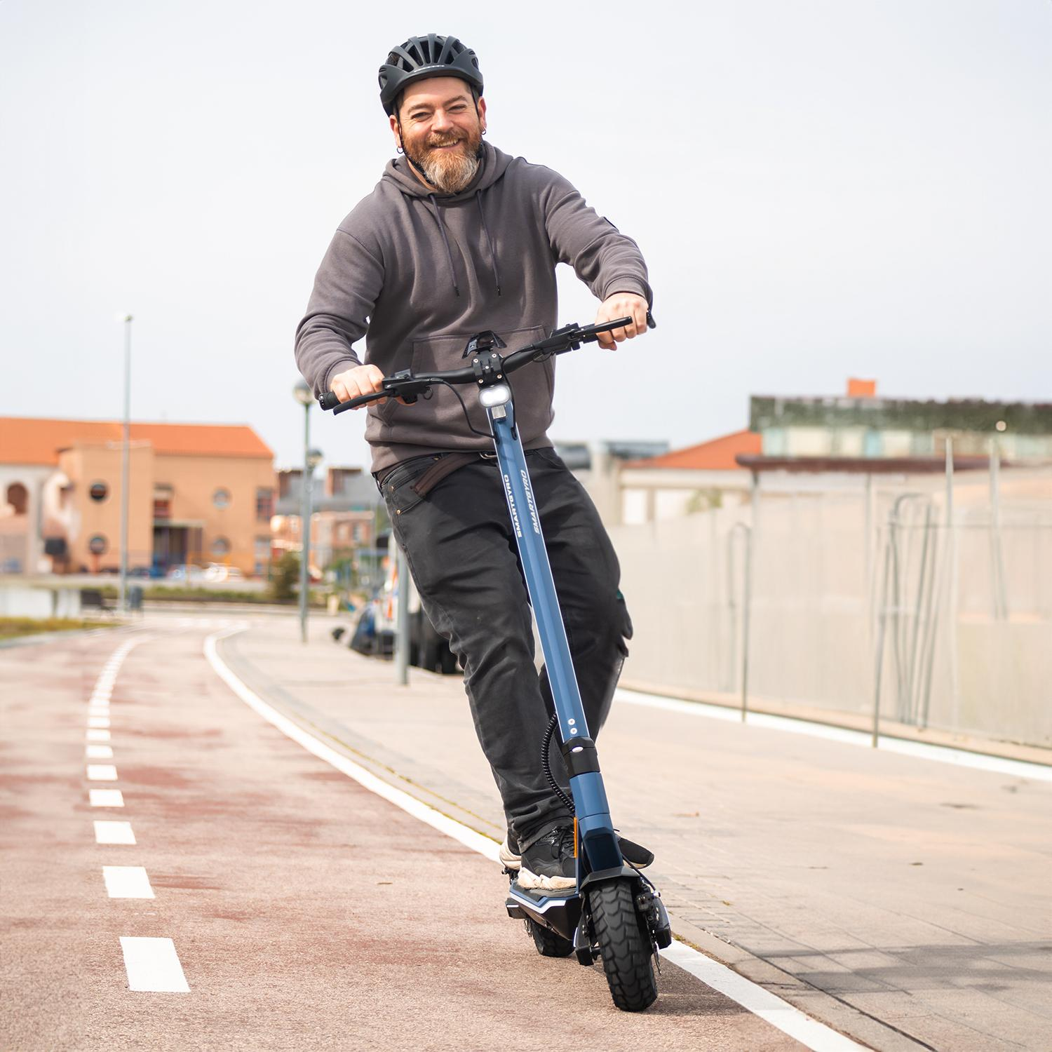 Un hombre anda en un scooter eléctrico en un carril bici, con casco y sudadera gris. Edificios al fondo.