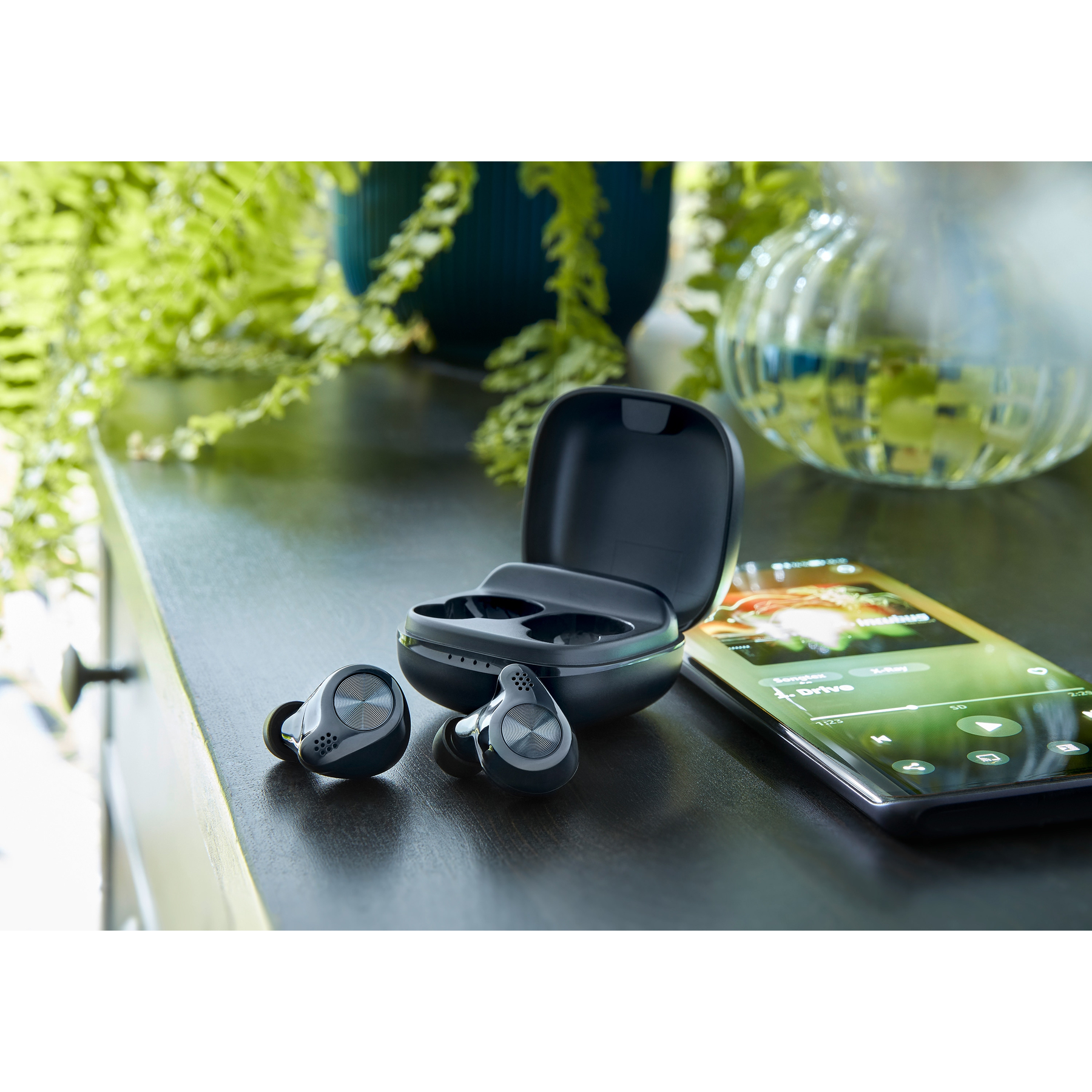 Auricolari wireless neri, custodia di ricarica, smartphone con musica e piante su superficie scura.