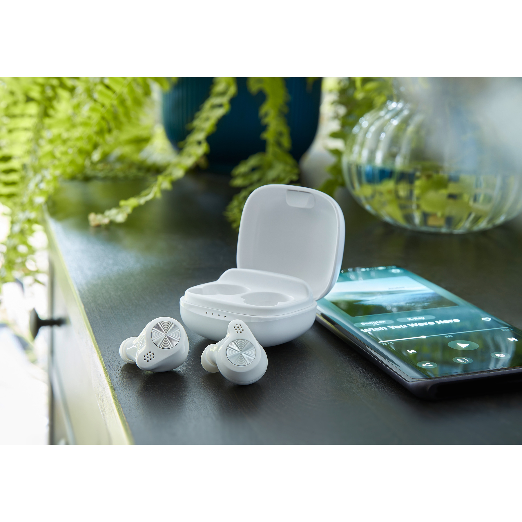 Weiße Ohrhörer und Ladecase auf dunkler Oberfläche neben einem Handy, das Musik abspielt. Grüne Pflanzen im Hintergrund.