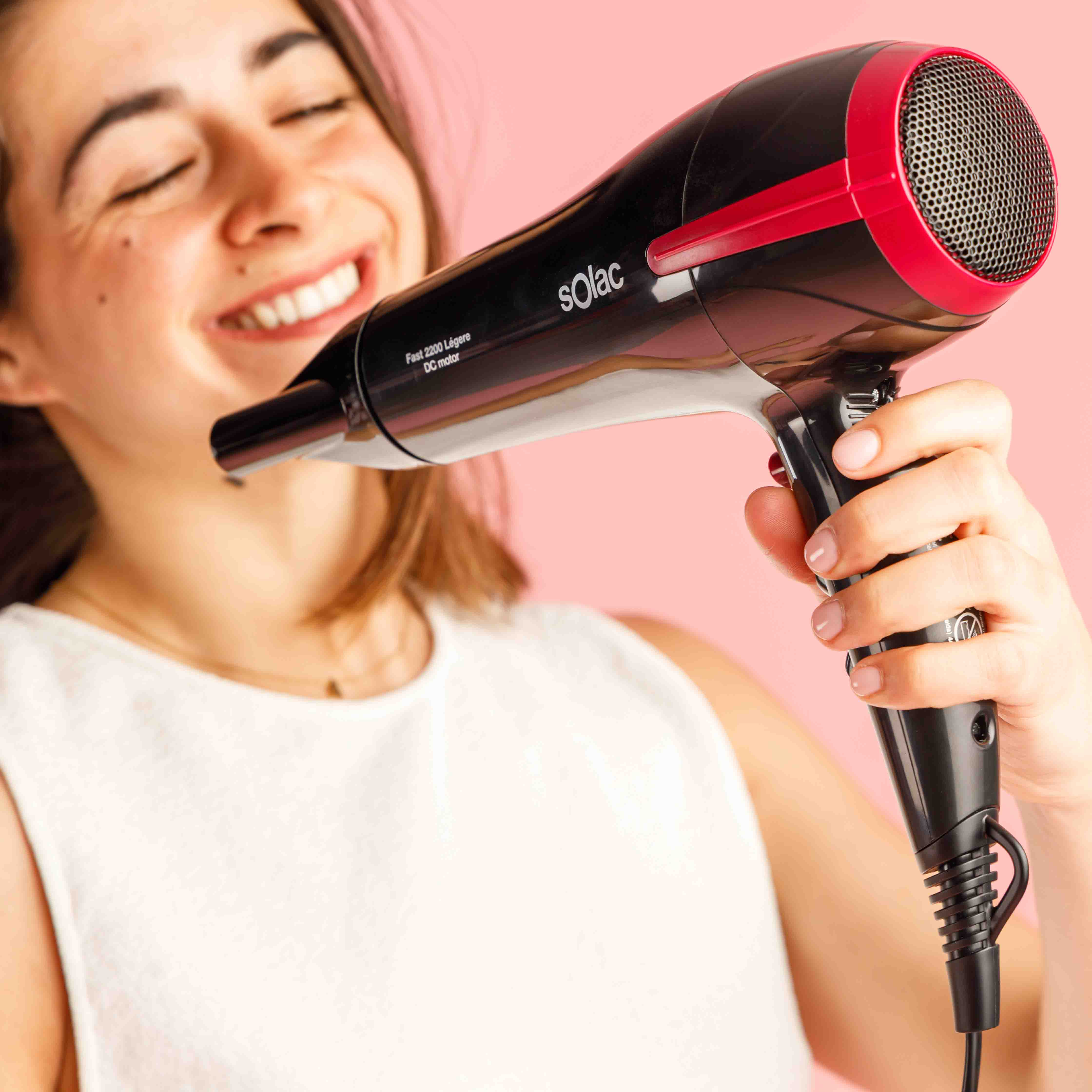 Kobieta trzyma czarno-czerwoną suszarkę do włosów, uśmiechając się z zamkniętymi oczami. Różowe tło.