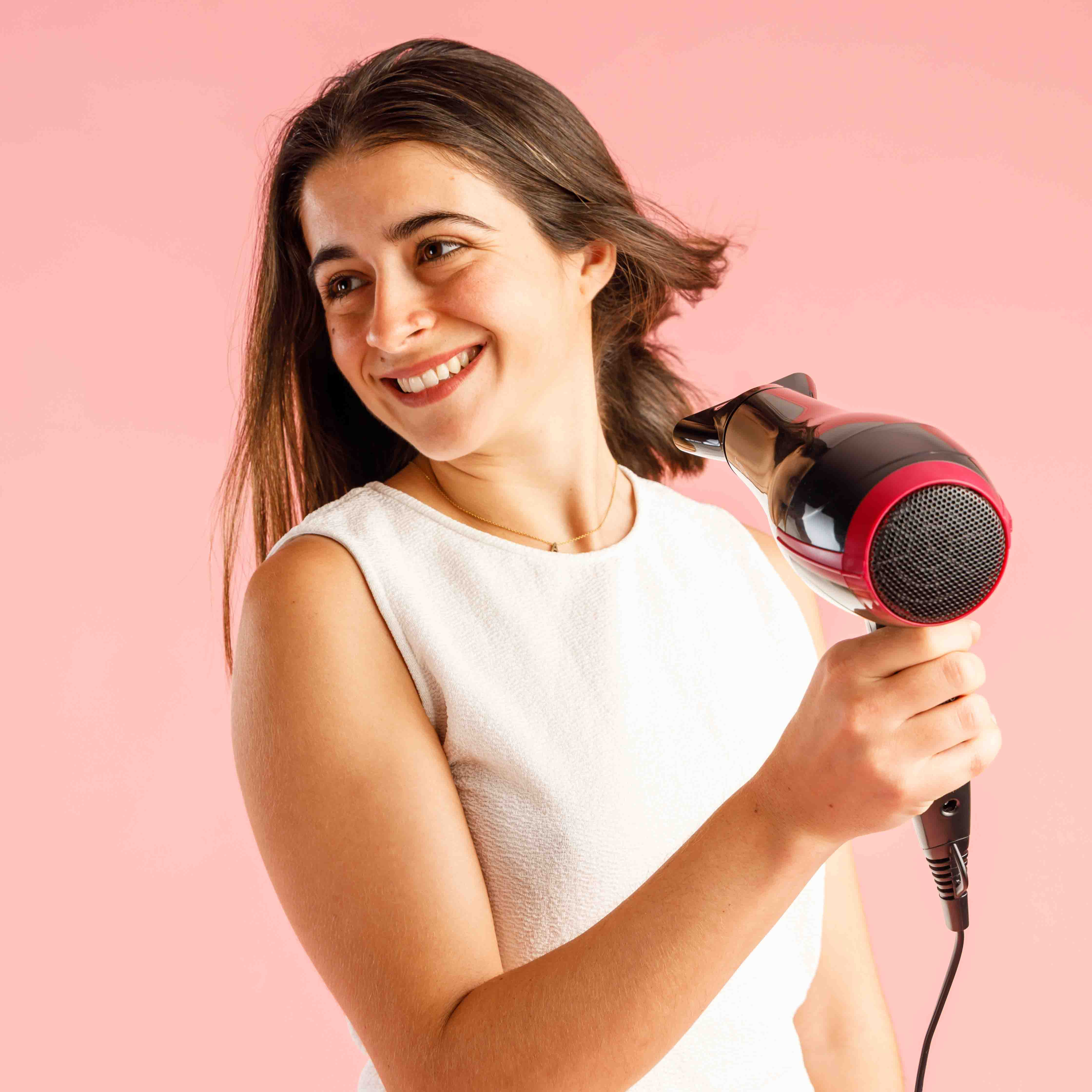 Uśmiechnięta kobieta suszy włosy suszarką. Różowe tło. Ma na sobie biały top i złoty naszyjnik.