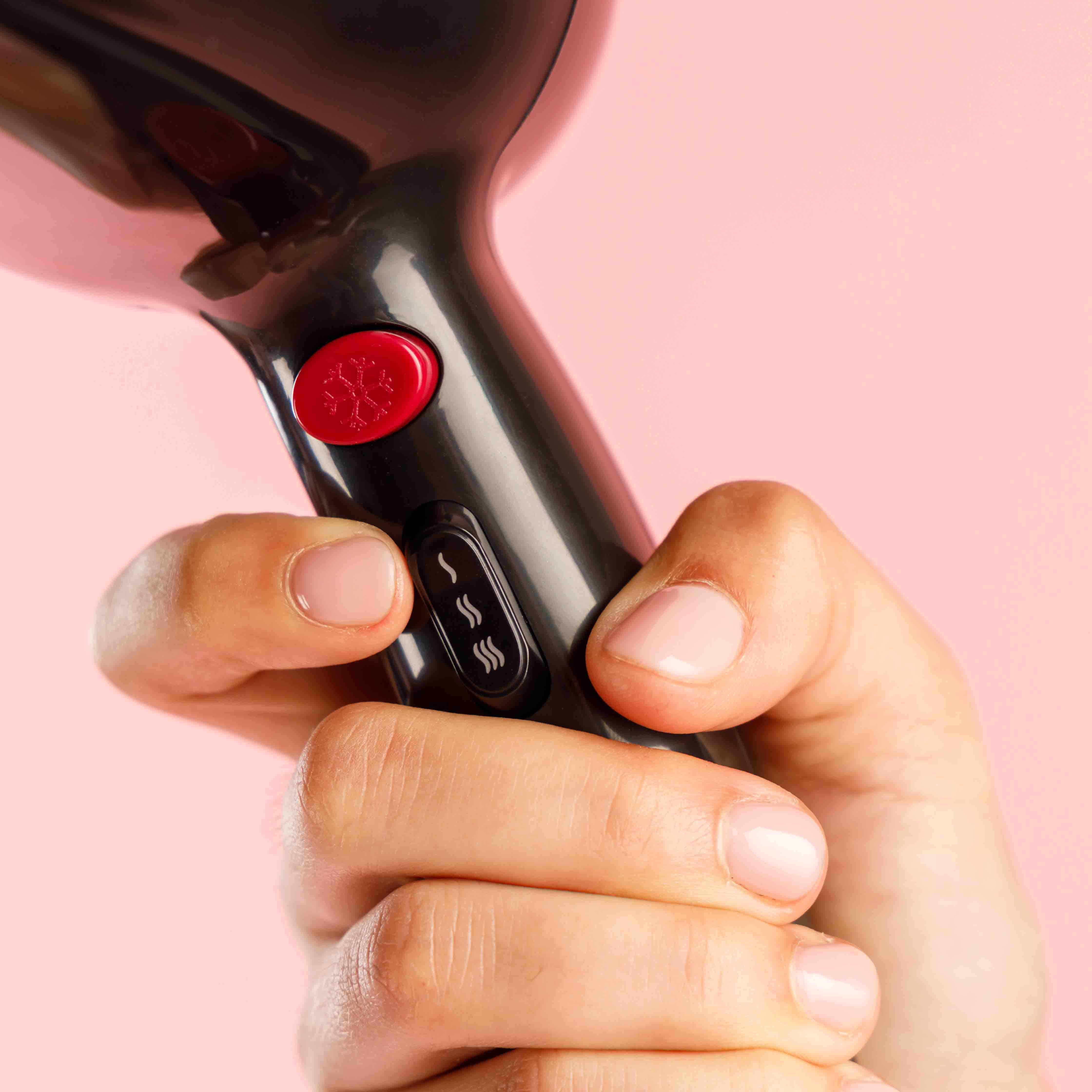 Dłoń trzymająca czarną suszarkę do włosów z czerwonym przyciskiem na różowym tle. Dłoń ma pomalowane paznokcie.