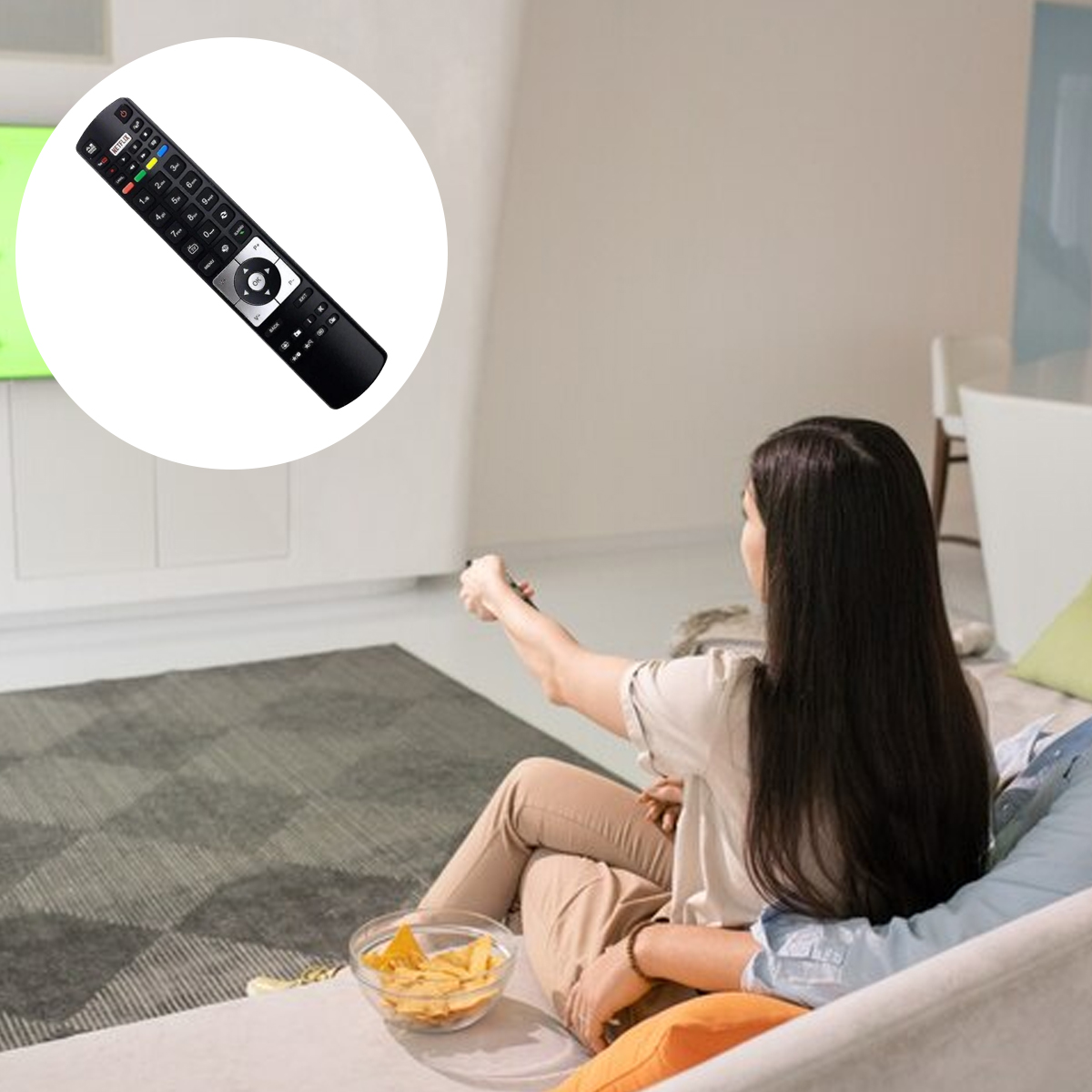 Eine Frau sitzt auf einem Sofa und hält eine Fernbedienung. Eine Schüssel Chips steht auf dem Sofa. Der Fernseher ist im Hintergrund.