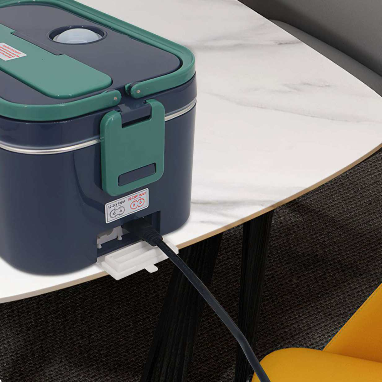 Eine blau-grüne Lunchbox mit eingestecktem Kabel, auf einem weißen Tisch neben einem Stuhl.