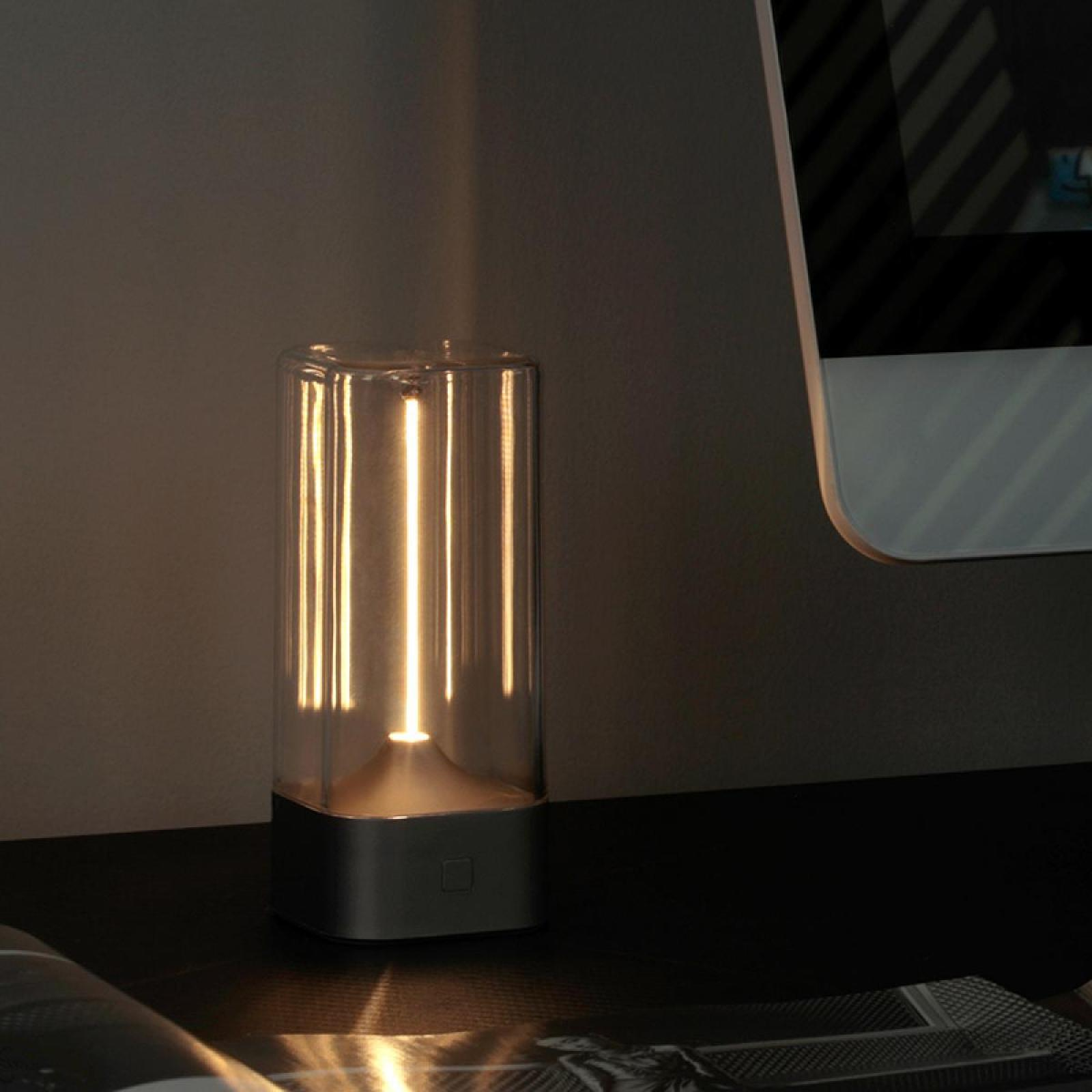 Eine beleuchtete, zylindrische Lampe mit Glas außen steht auf einem dunklen Schreibtisch. Ein Computermonitor ist im Hintergrund sichtbar.