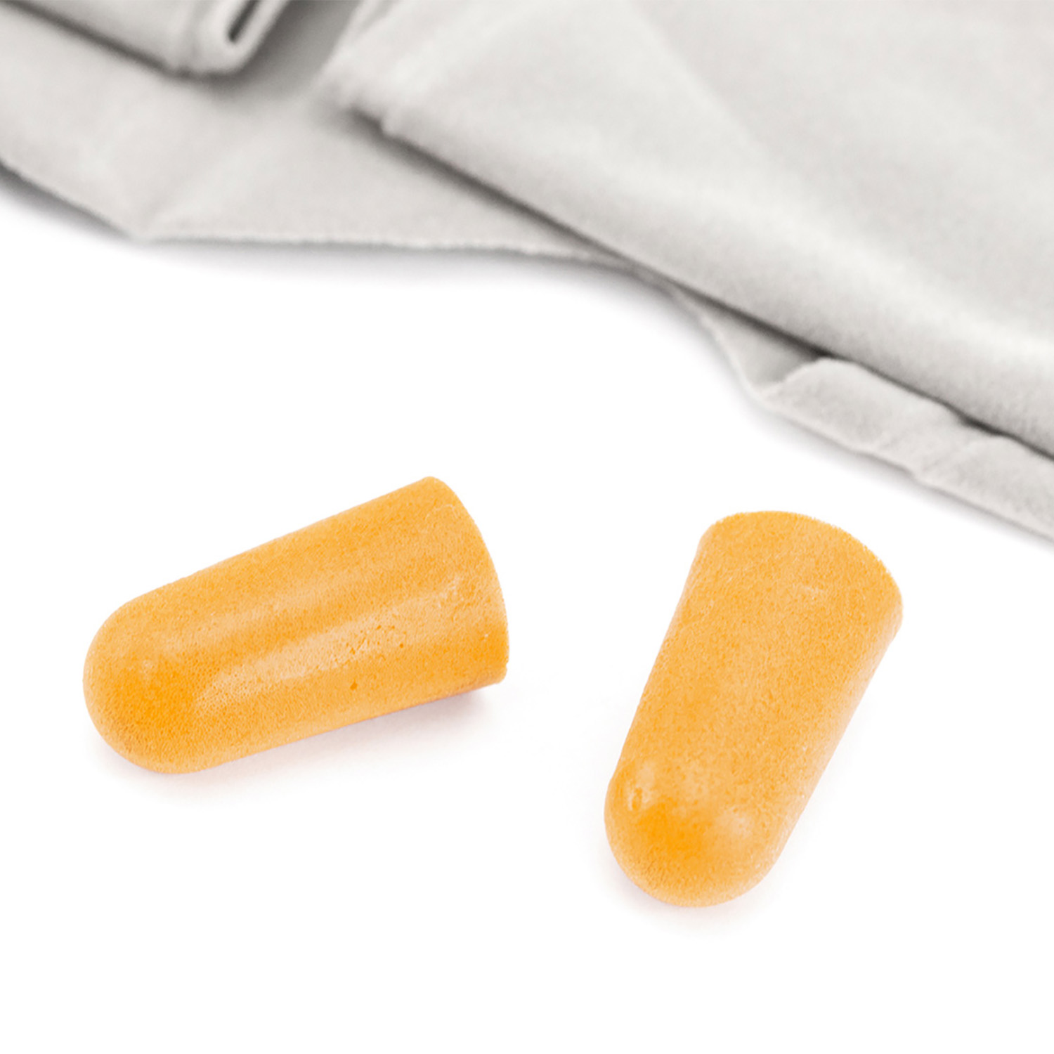 Dos tapones para los oídos naranjas sobre una superficie blanca, con un paño gris en el fondo. Los tapones son cilíndricos y de espuma.