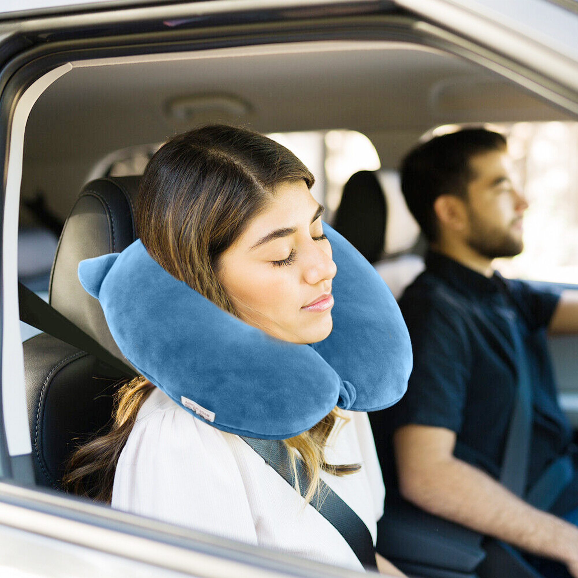 Una mujer duerme en un coche con una almohada azul en el cuello. Un hombre conduce detrás.