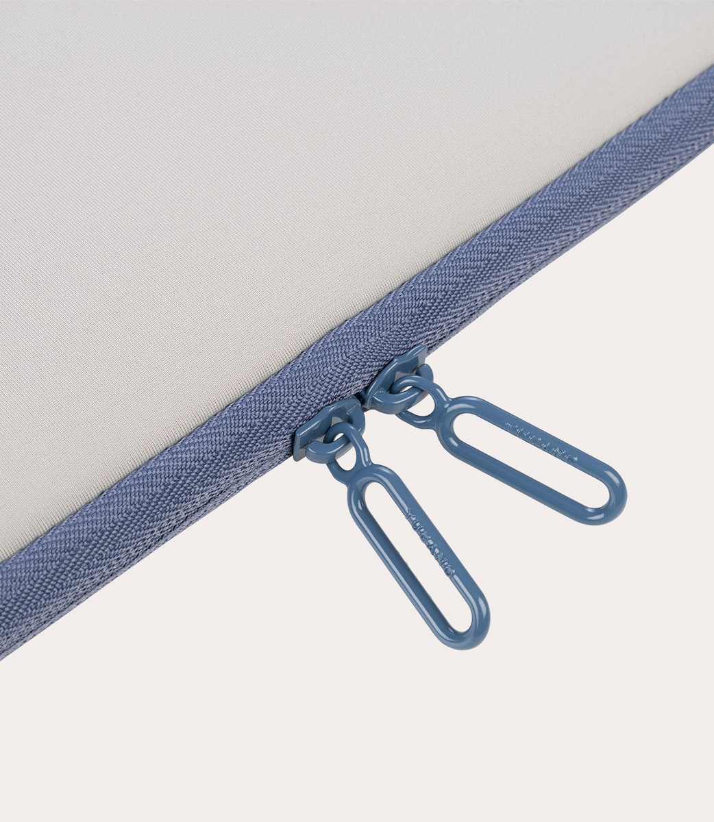 Nahaufnahme eines blauen Reißverschlusses auf hellem, grauem Stoff. Zwei passende blaue Reißverschlusszipper sind sichtbar, mit weißem Hintergrund.