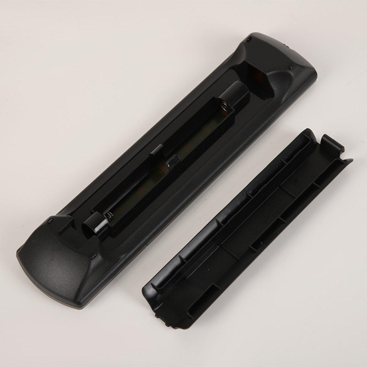 Eine schwarze Fernbedienung mit geöffnetem Batteriefach liegt auf einer weißen Oberfläche. Die Fachabdeckung ist separat.
