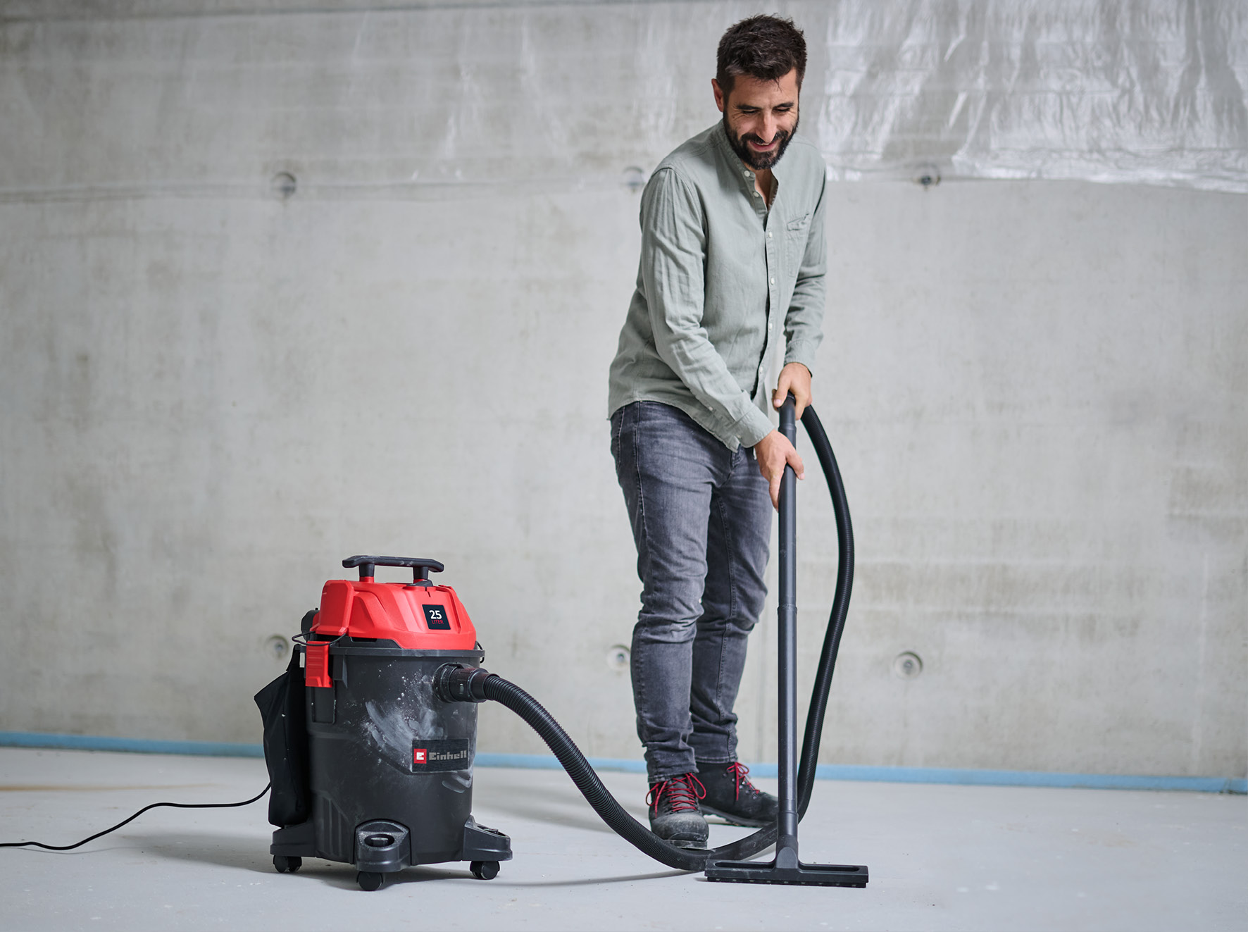 Ein Mann saugt einen Betonboden mit einem schwarz-roten Staubsauger. Er trägt ein hellgrünes Hemd und Jeans.