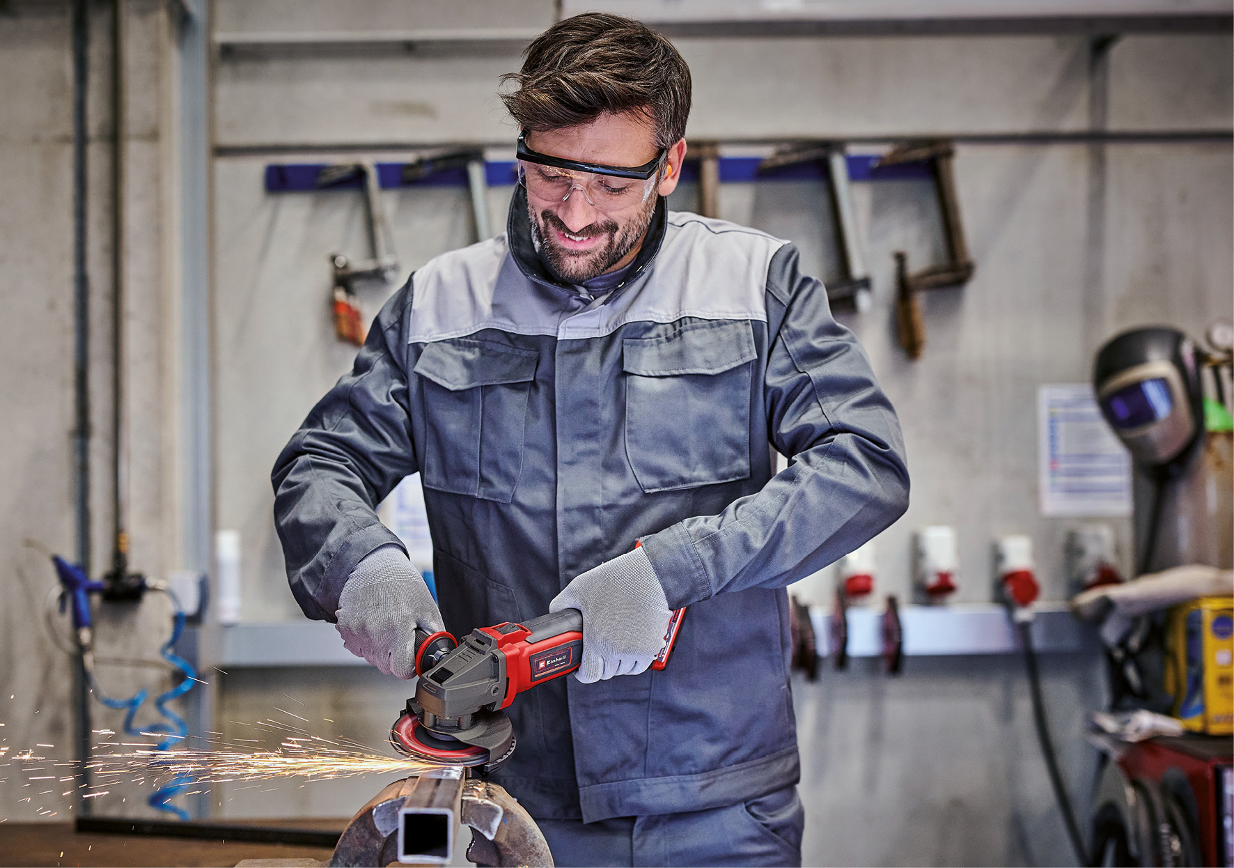 Ein Mann schleift Metall mit einem roten Schleifer, Funken fliegen. Er trägt eine Schutzbrille und Arbeitskleidung. Werkstatthintergrund.