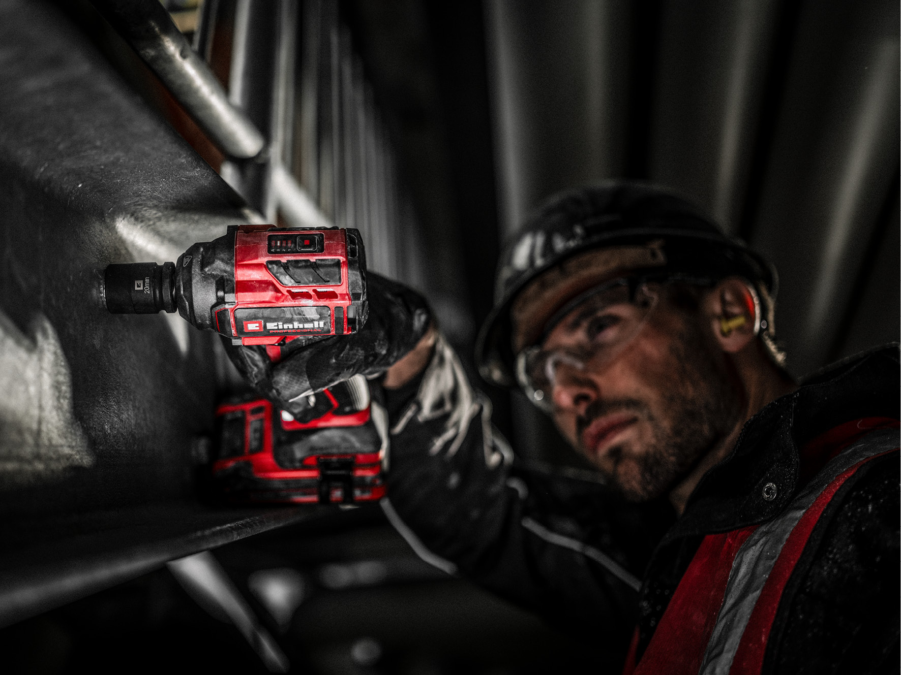 Ein Arbeiter in Schutzkleidung verwendet ein rotes Elektrowerkzeug. Der Hintergrund ist dunkel mit Metallstrukturen, Fokus auf Werkzeug und Arbeiter.