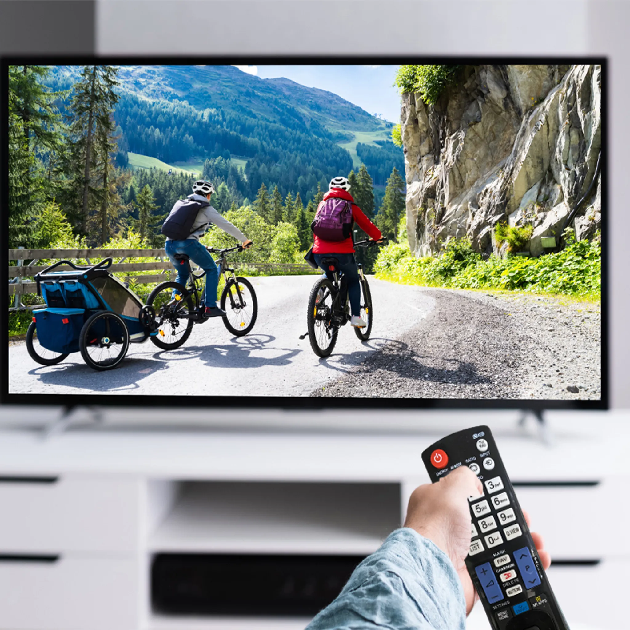 Ein Fernseher zeigt zwei Radfahrer auf einer Bergstraße. Eine Person hält eine Fernbedienung. Weißer Schrank im Vordergrund.