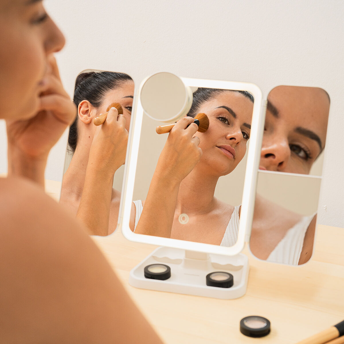 Una donna si trucca davanti a uno specchio illuminato. Lo specchio riflette il suo viso e le sue mani. Trucco e pennelli sono sul tavolo.