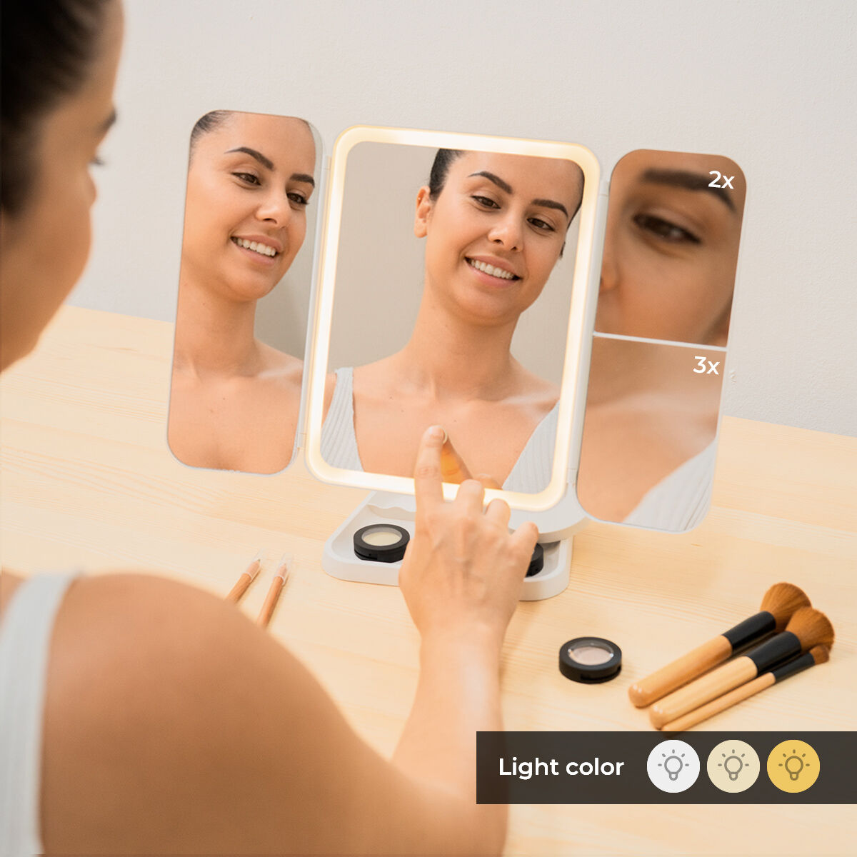 Una donna usa uno specchio per il trucco illuminato. Lo specchio ha tre pannelli, quello centrale è illuminato. Pennelli e prodotti per il trucco sono visibili sul tavolo.