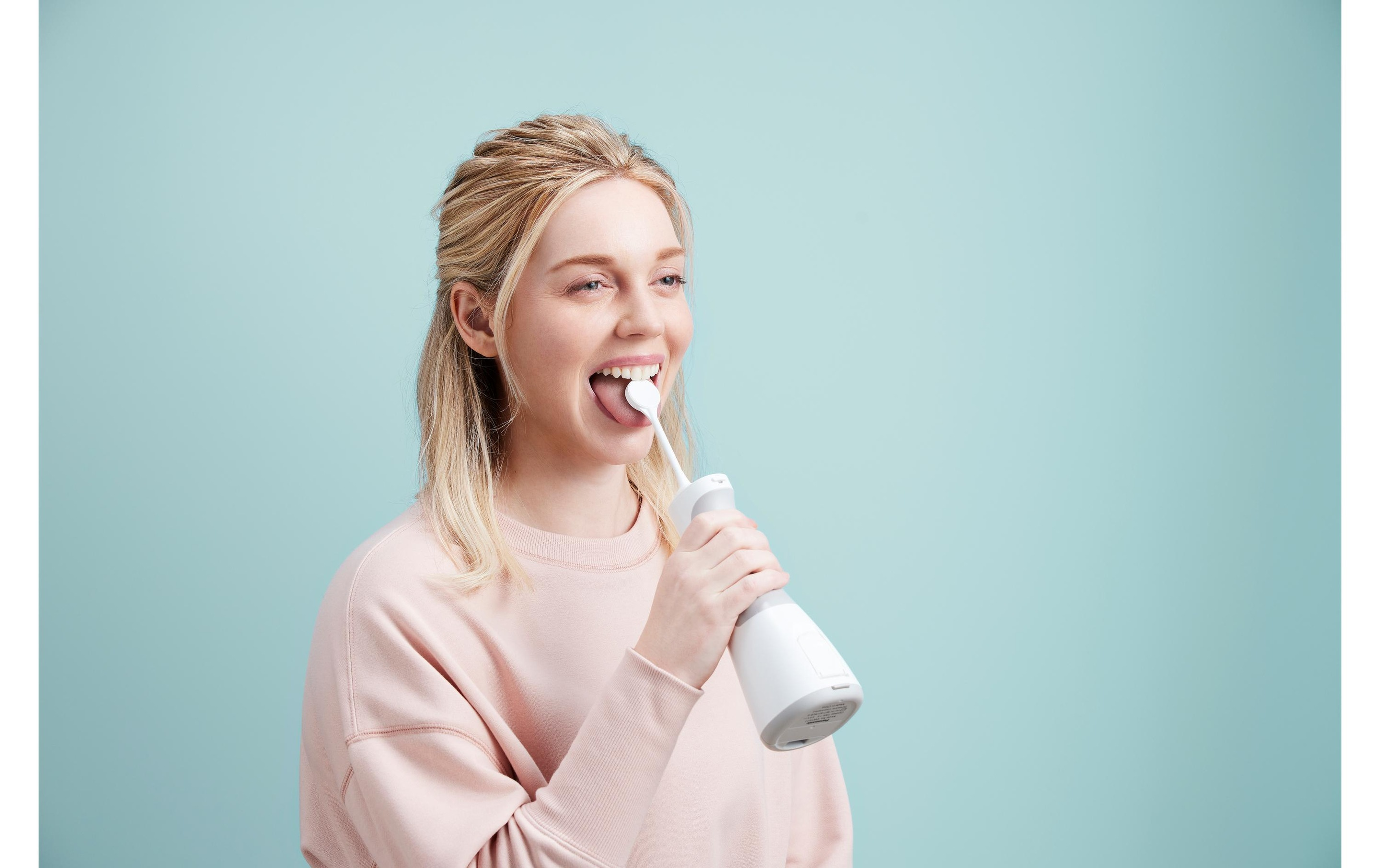 Eine Frau benutzt einen Munddusche. Sie trägt einen rosa Pullover. Der Hintergrund ist hellblau.