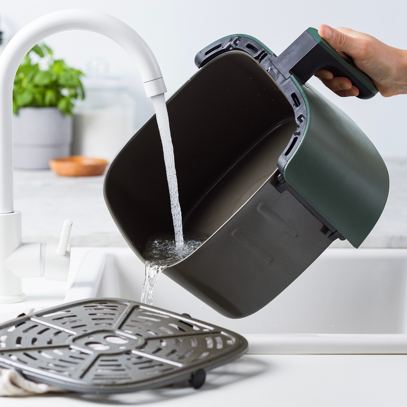 Eine Person wäscht einen Airfryer-Korb unter einem Wasserhahn. Wasser fließt über ein Gestell in den dunkelgrünen Korb.