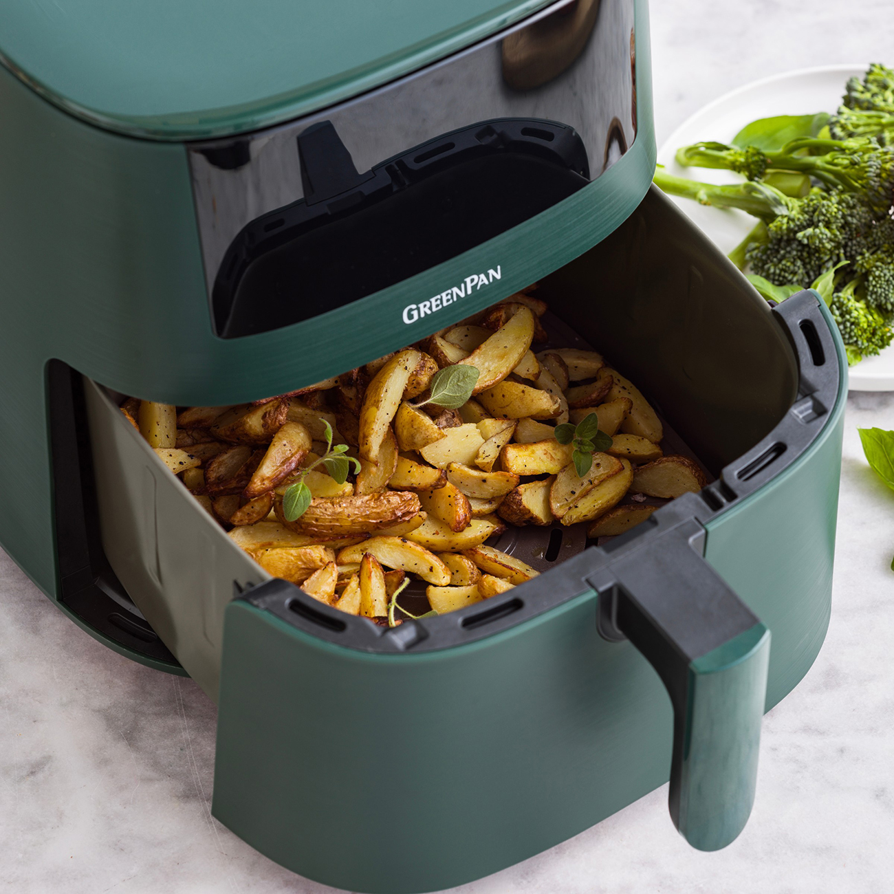 Ein Airfryer mit gekochten Kartoffelspalten. Brokkoli und Basilikum liegen auf einem weißen Teller. Der Airfryer ist grün.