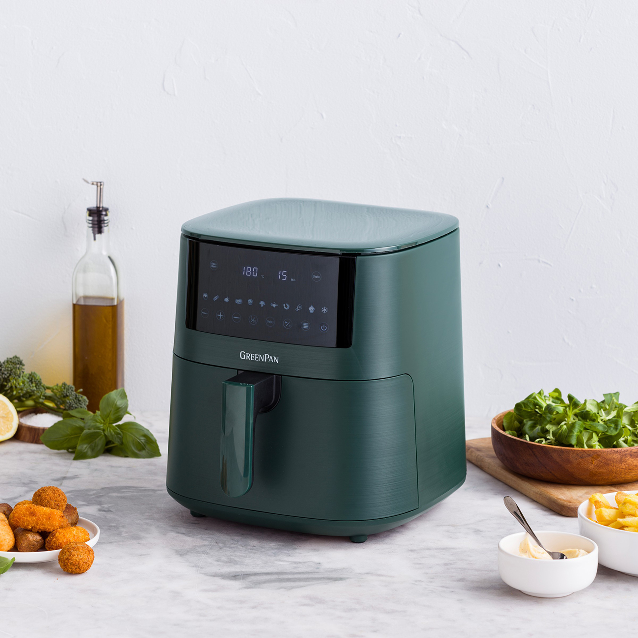 Ein grüner Airfryer steht auf einer Marmoroberfläche. Drumherum befinden sich Speisen wie Pommes, Salat und ein Teller mit Vorspeisen.