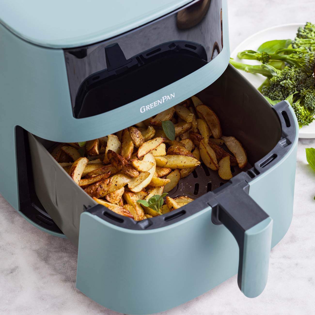Eine Heißluftfritteuse mit gekochten Kartoffelspalten. GreenPan-Logo auf dem Gerät. Brokkoli und Basilikumblätter im Hintergrund.
