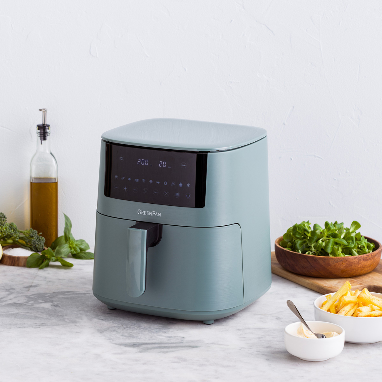 Ein tealfarbener Luftfritteuse steht auf einer Marmortheke neben Pommes Frites, Grünzeug und Olivenöl. Das Display zeigt 200 Grad und 20 Minuten.