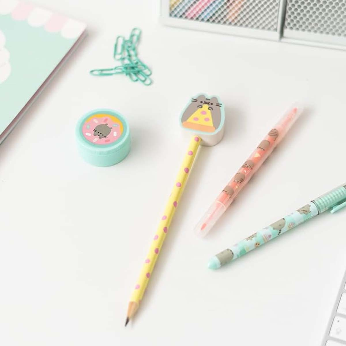 Ein weißer Schreibtisch mit Schreibwaren. Enthält einen Bleistift mit rosa Punkten, Stifte, einen donutformigen Radiergummi und Büroklammern. Weiche Farben.
