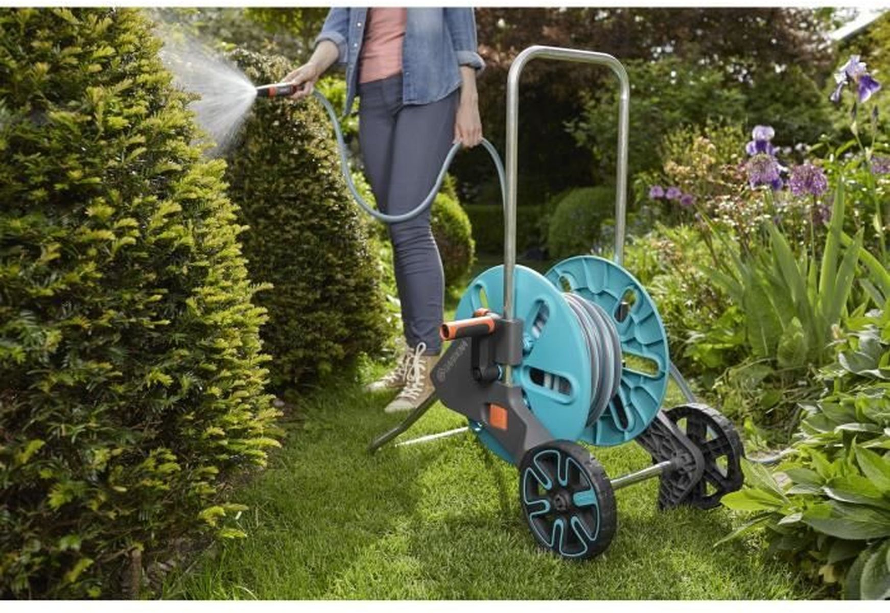Una persona innaffia un cespuglio verde con un tubo flessibile in un giardino. Un avvolgitubo è sulla destra. Altre piante e fiori sono sullo sfondo.