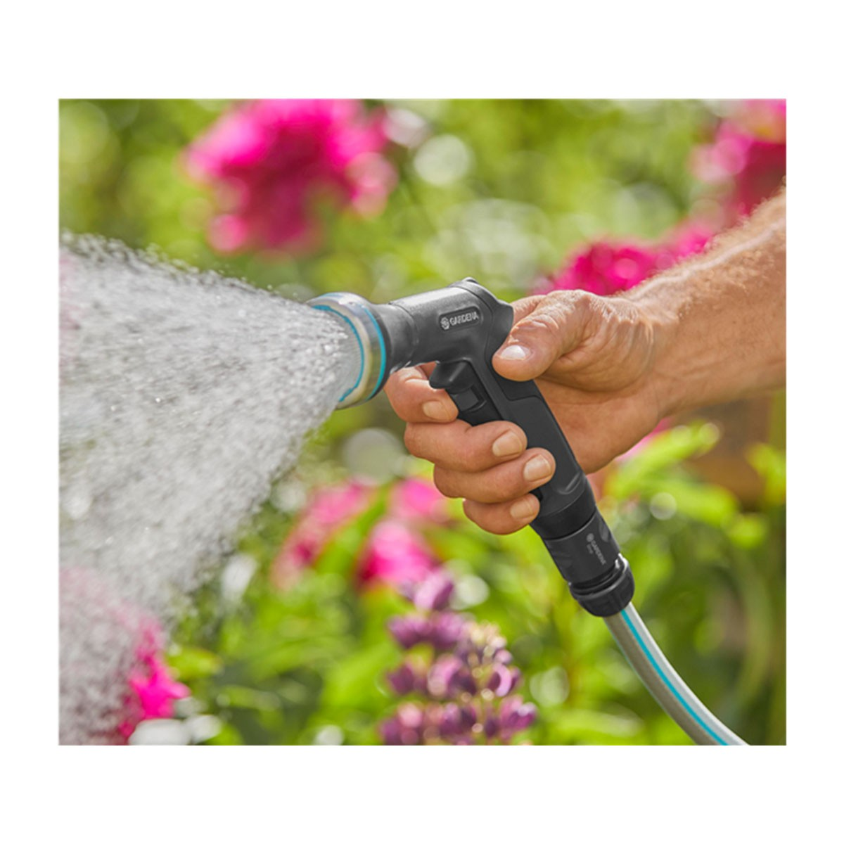 Una mano tiene un ugello per tubo dell'acqua Gardena nero e grigio, che spruzza acqua. Fiori rosa e fogliame verde sullo sfondo.
