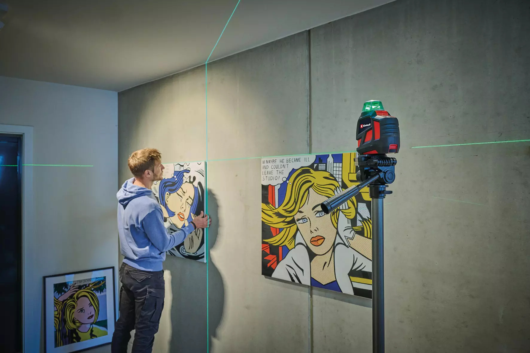 Mężczyzna wiesza dzieła sztuki za pomocą poziomicy laserowej. Portrety kobiet w stylu pop-art na ścianie. Poziomica laserowa na statywie.