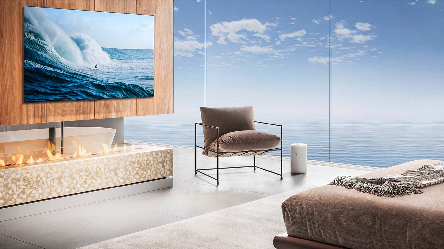 Un soggiorno presenta una TV che mostra un surfista sull'onda. Un camino brilla sotto. Una sedia è di fronte alla vista mare.