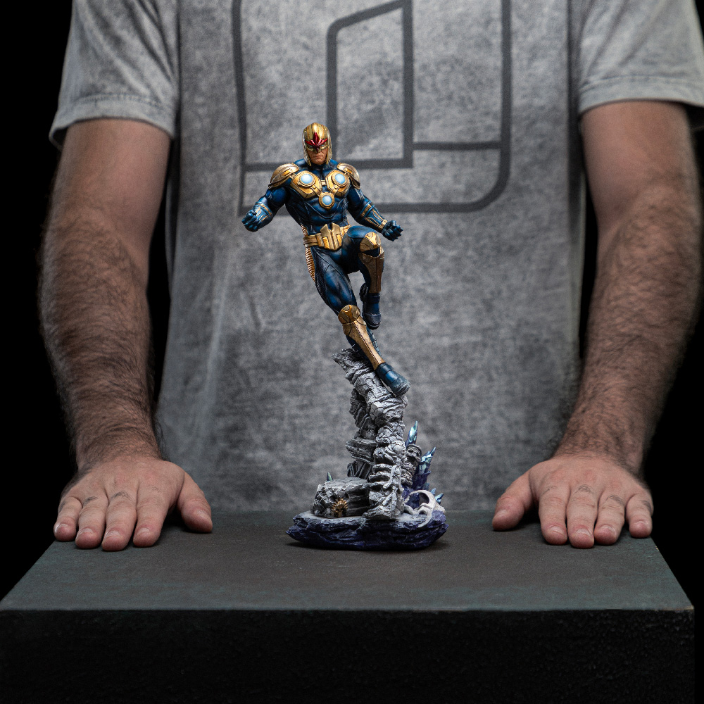 Una persona tiene in mano una statua di Nova. La statua è di un supereroe in tuta blu e oro, che salta da una base rocciosa.