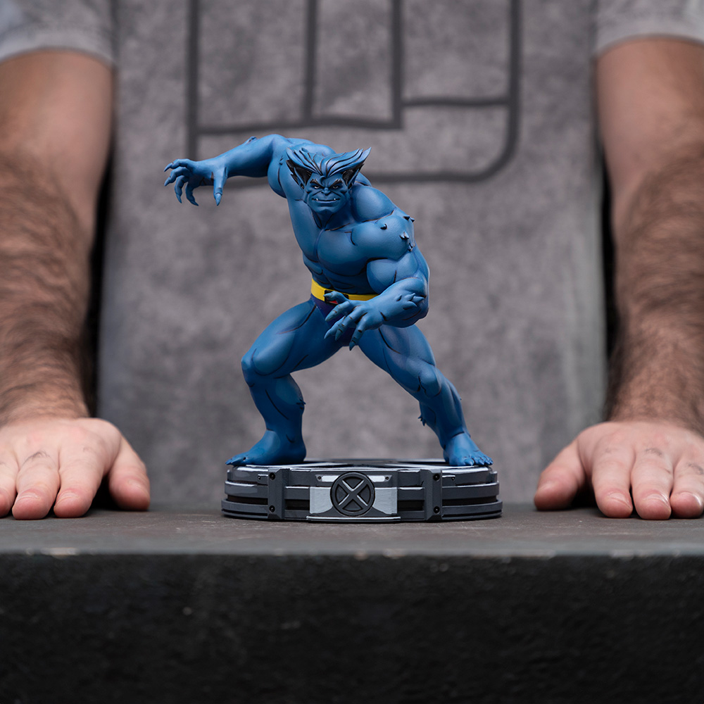 Eine blaue Beast-Figur mit gelbem Gürtel steht auf einem Ständer. Die Hände der Person sind an den Seiten. Der Hintergrund ist grau.