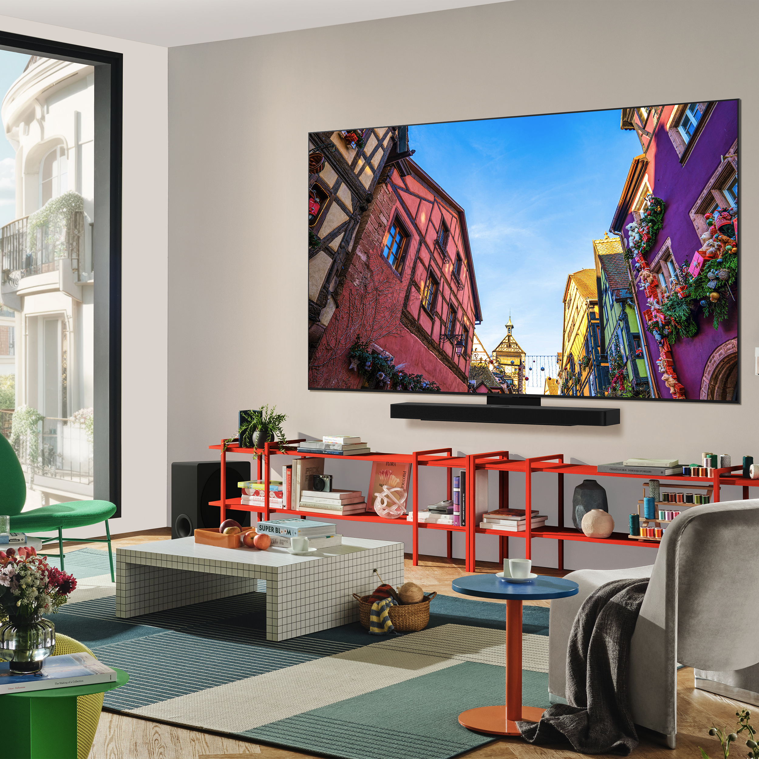 Salon avec grand téléviseur, vue de bâtiments colorés à l'écran, étagères rouges, table blanche et chaise verte.