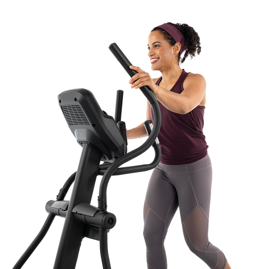 Una mujer sonríe mientras usa una máquina elíptica. Viste ropa deportiva, incluyendo una camiseta burdeos y mallas grises.