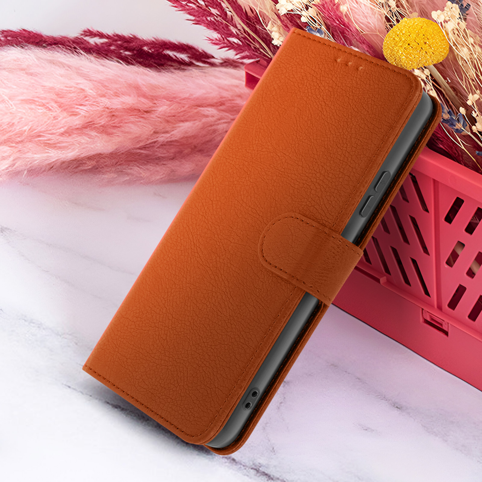 Un étui de téléphone en cuir orange est ouvert, posé sur une surface en marbre. Fleurs séchées roses et jaunes en arrière-plan.