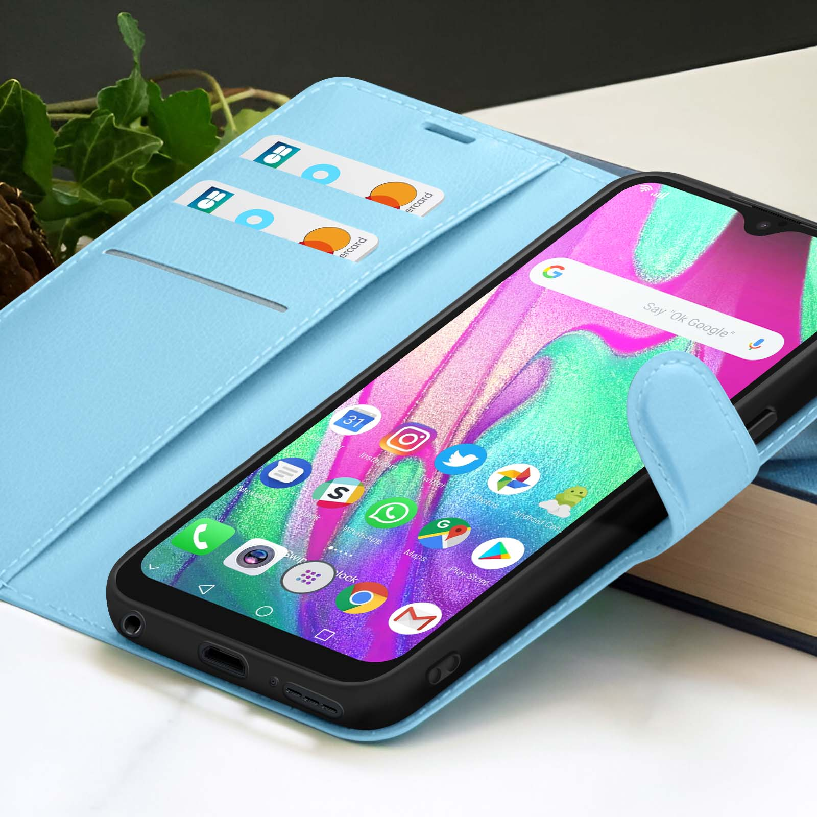 Un smartphone dans un étui bleu clair affichant des icônes d'applications. Cartes de crédit dans l'étui. Arrière-plan: livre et feuillage.