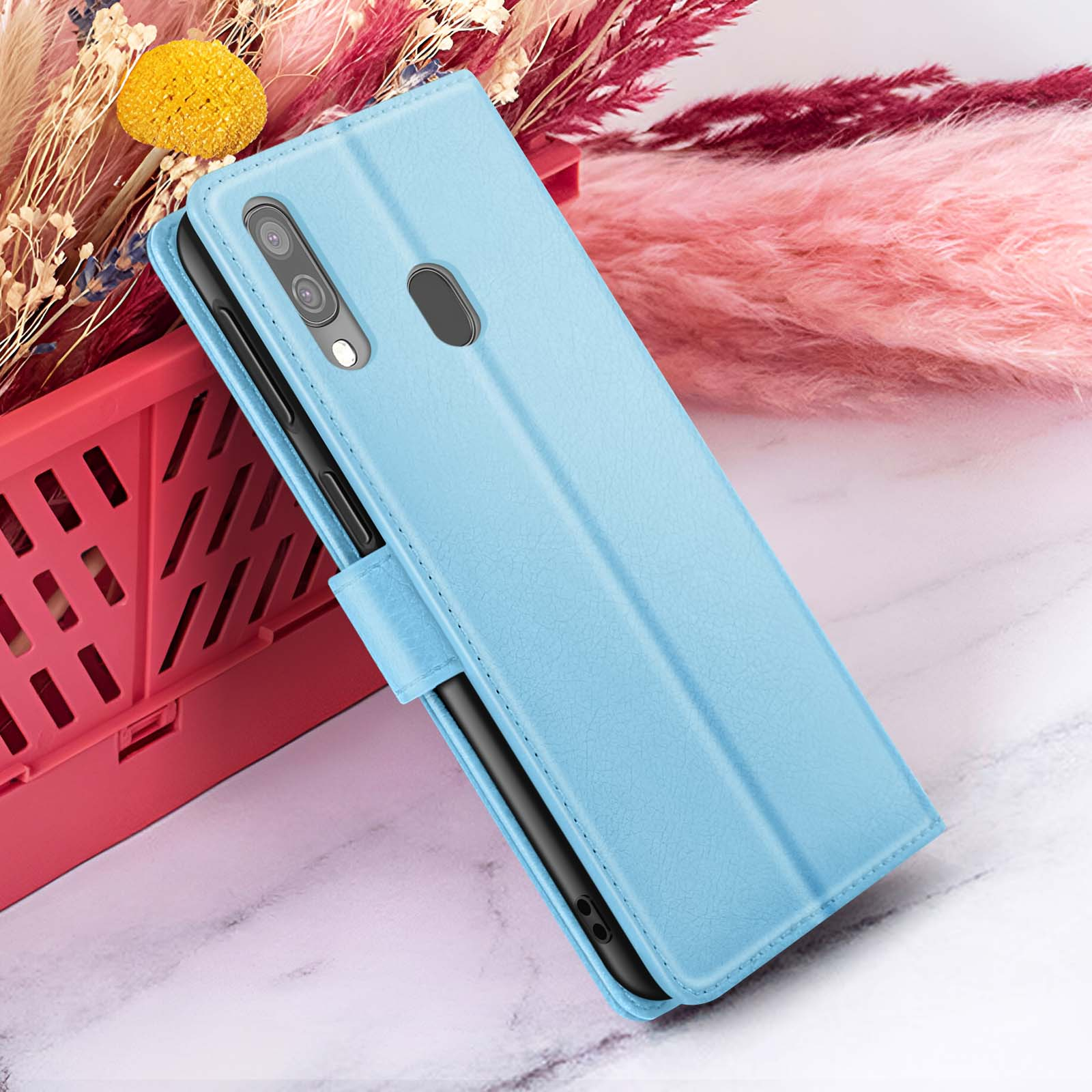 Un étui de téléphone bleu clair est ouvert, révélant un téléphone noir. Des fleurs et un récipient rouge en arrière-plan.