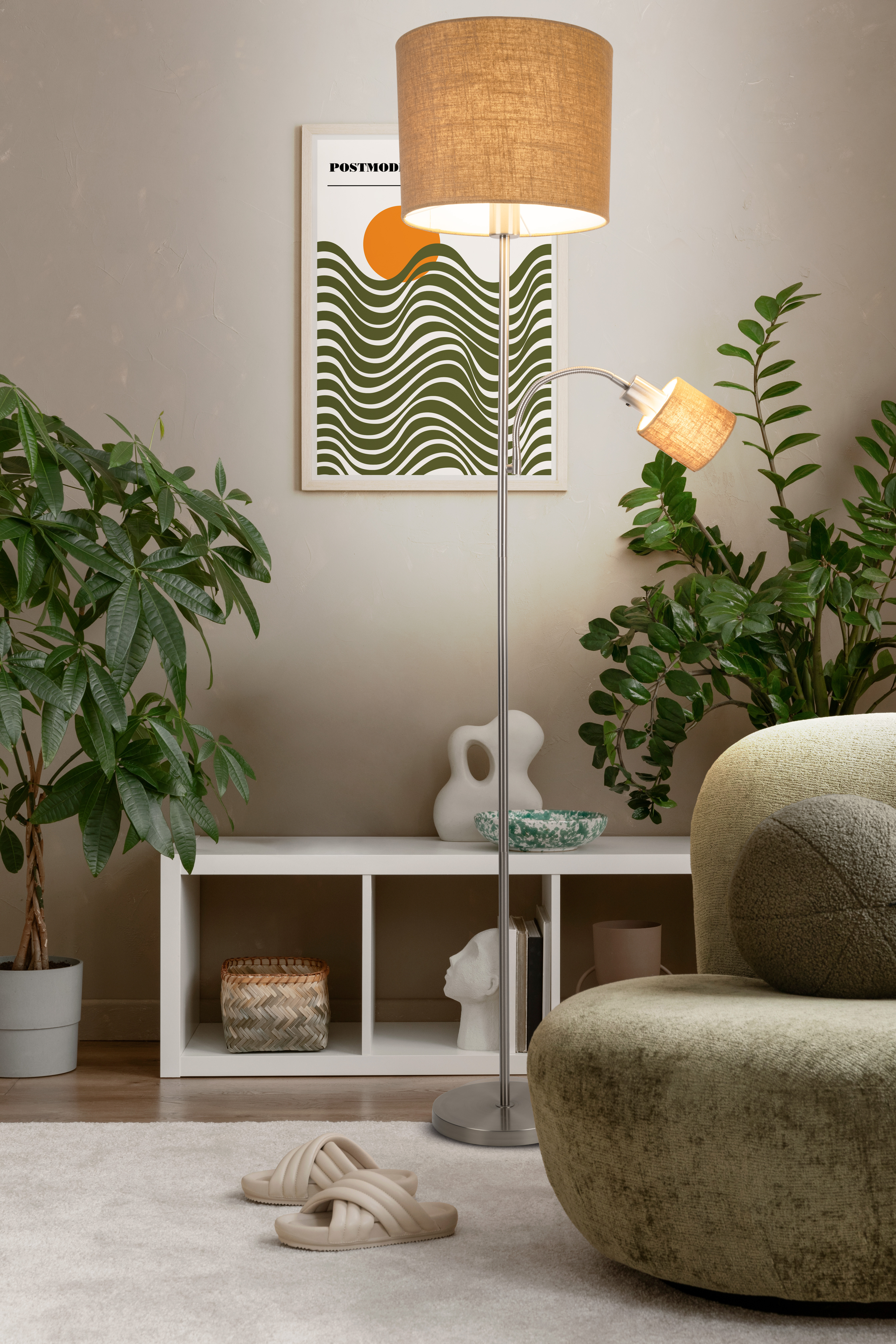 Ein Zimmer mit Stehlampe, Pflanzen, weißem Regal mit Deko und olivgrünem Sessel. Beige Hausschuhe liegen auf dem Teppich.