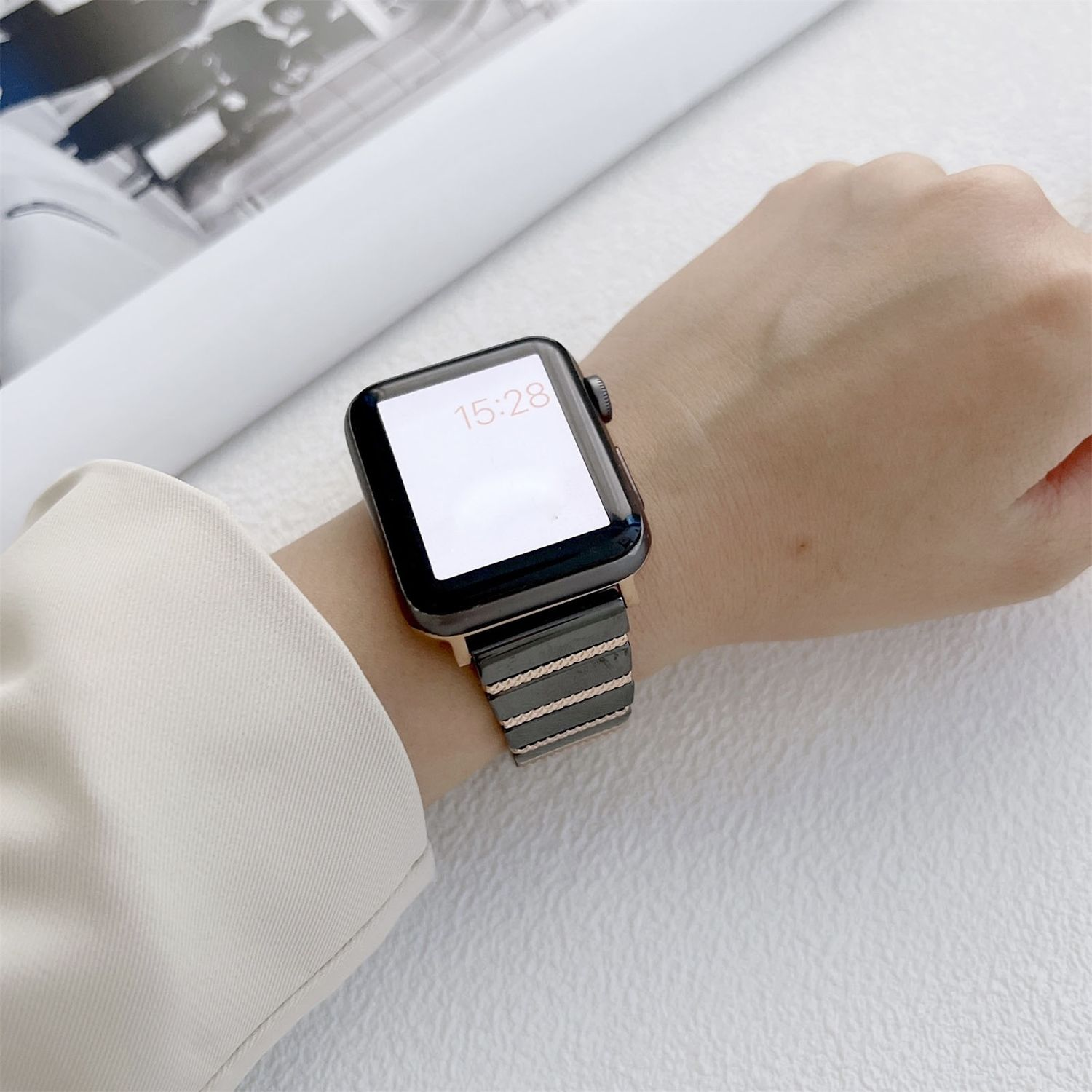 Handgelenk mit Smartwatch, die 15:28 anzeigt. Die Uhr hat einen schwarzen Bildschirm und ein graues Armband mit goldenen Akzenten. Der Hintergrund ist weiß.