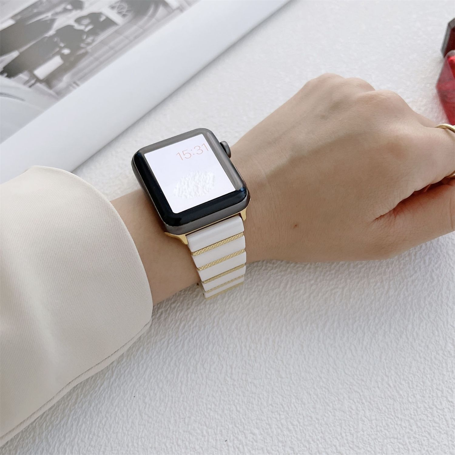 Hand mit weißer und goldener Uhr. Die Uhr zeigt 15:31. Ein cremefarbenes Hemd und ein weißer Hintergrund sind ebenfalls sichtbar.