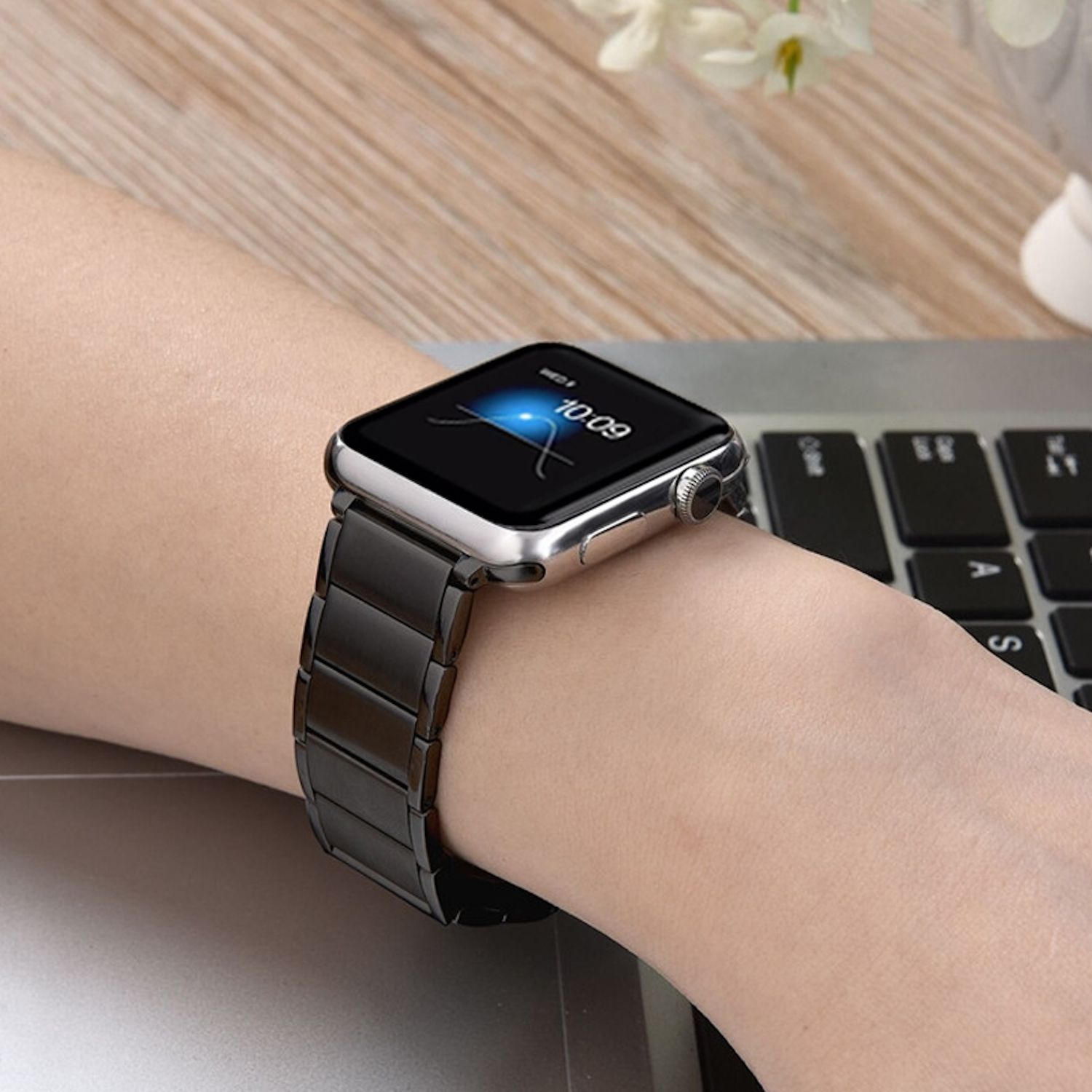 Ein Handgelenk mit einer schwarzen Metall-Apple Watch. Die Uhr zeigt 10:09 Uhr an. Eine Laptop-Tastatur und ein Holztisch sind im Hintergrund.