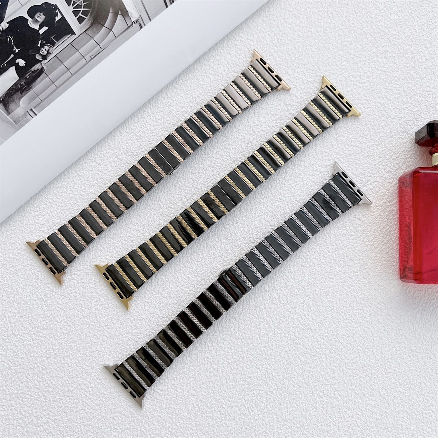 Drei Uhrenarmbänder mit schwarzen, goldenen und silbernen Gliedern auf einer weißen, strukturierten Oberfläche, mit Parfümflasche und SW-Bild im Hintergrund.